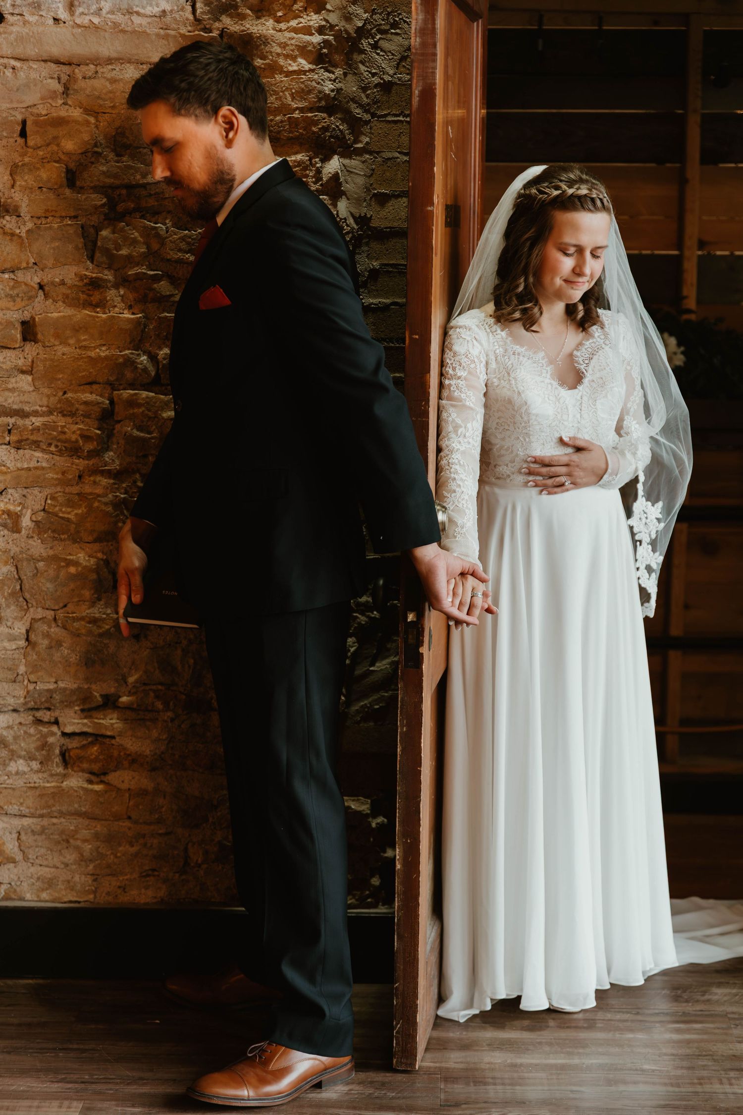 Wedding couple in formal attire standing against exposed brick wall under crystal chandeliers.