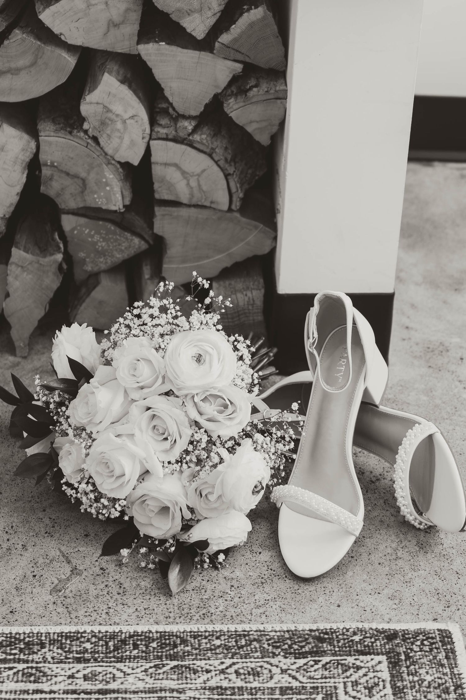 White bridal shoes and rose bouquet arranged on decorative rug with stacked firewood background.
