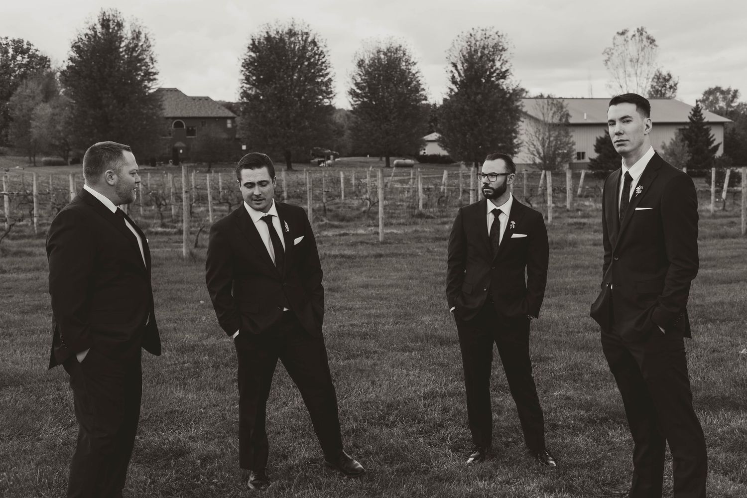 Groomsmen in dark suits standing together on grass at outdoor wedding venue.