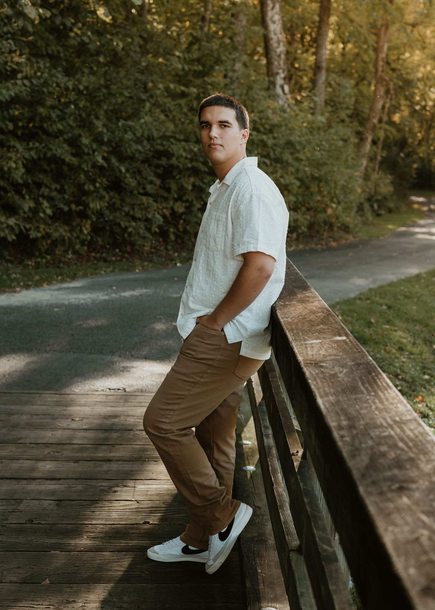 Young man in white shirt and khaki pants leaning on wooden bridge railing surrounded by lush green trees.