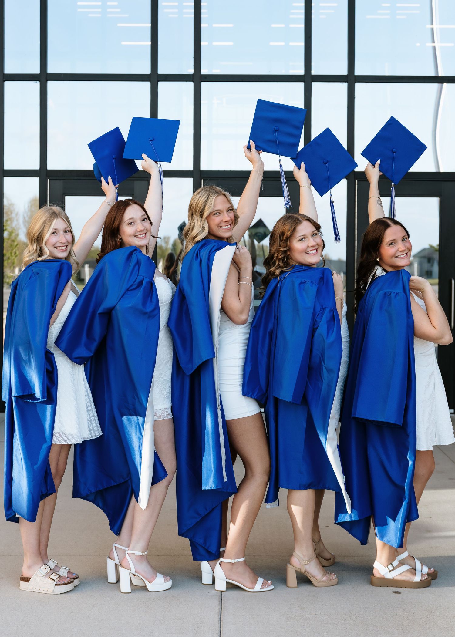Five female graduates in blue caps and gowns raising their mortarboards outdoors.