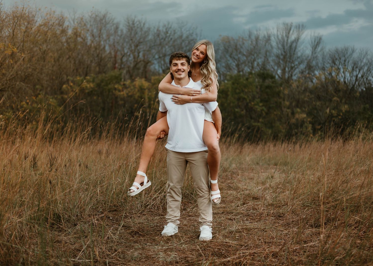 A couple shares a playful piggyback ride moment in a golden field with autumn trees in the background.