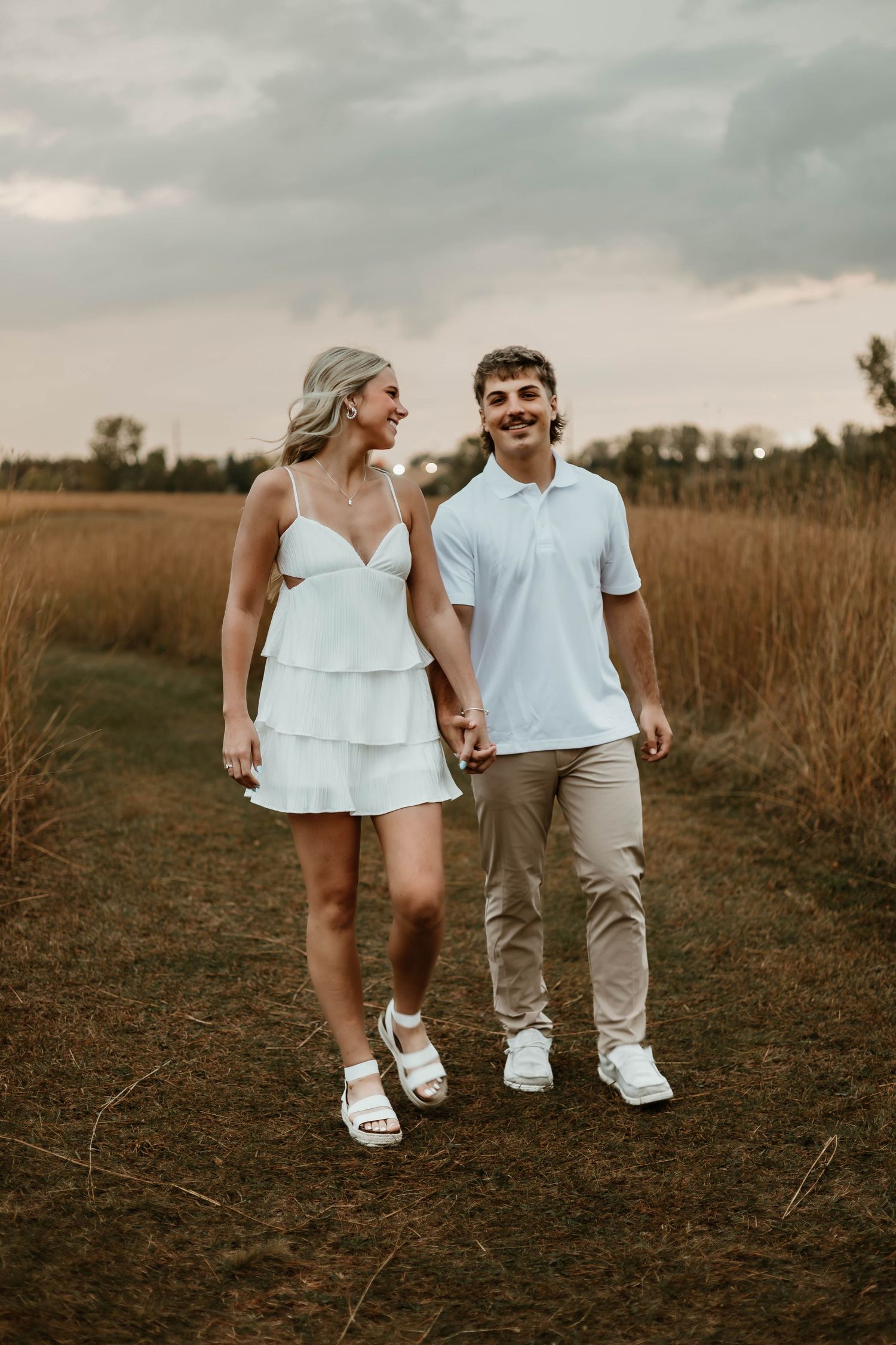 A couple walking together on a dirt path through tall golden grass at sunset.