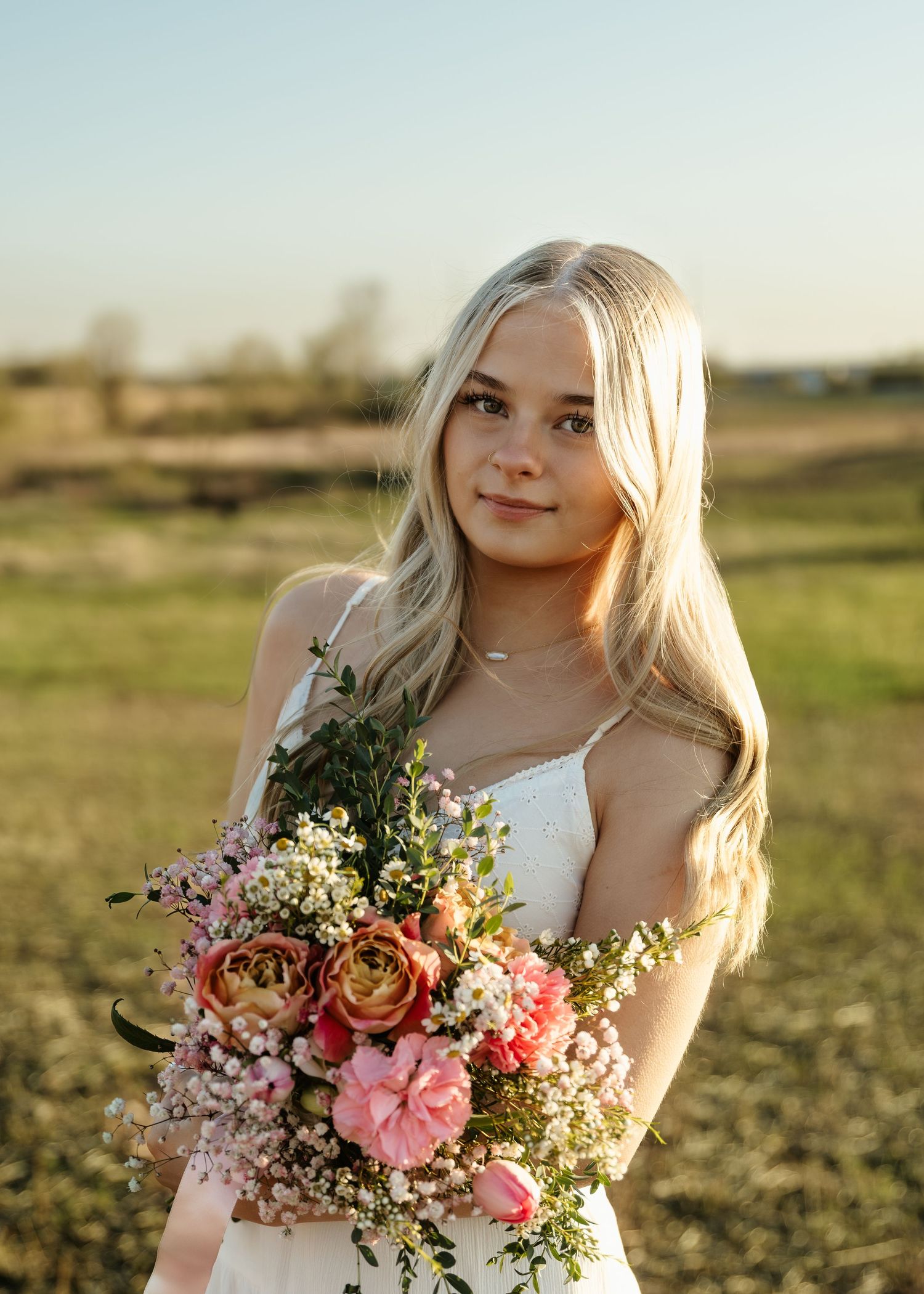 Young blonde woman in white dress holding a colorful bouquet of pink roses and wildflowers in a sunny outdoor field.