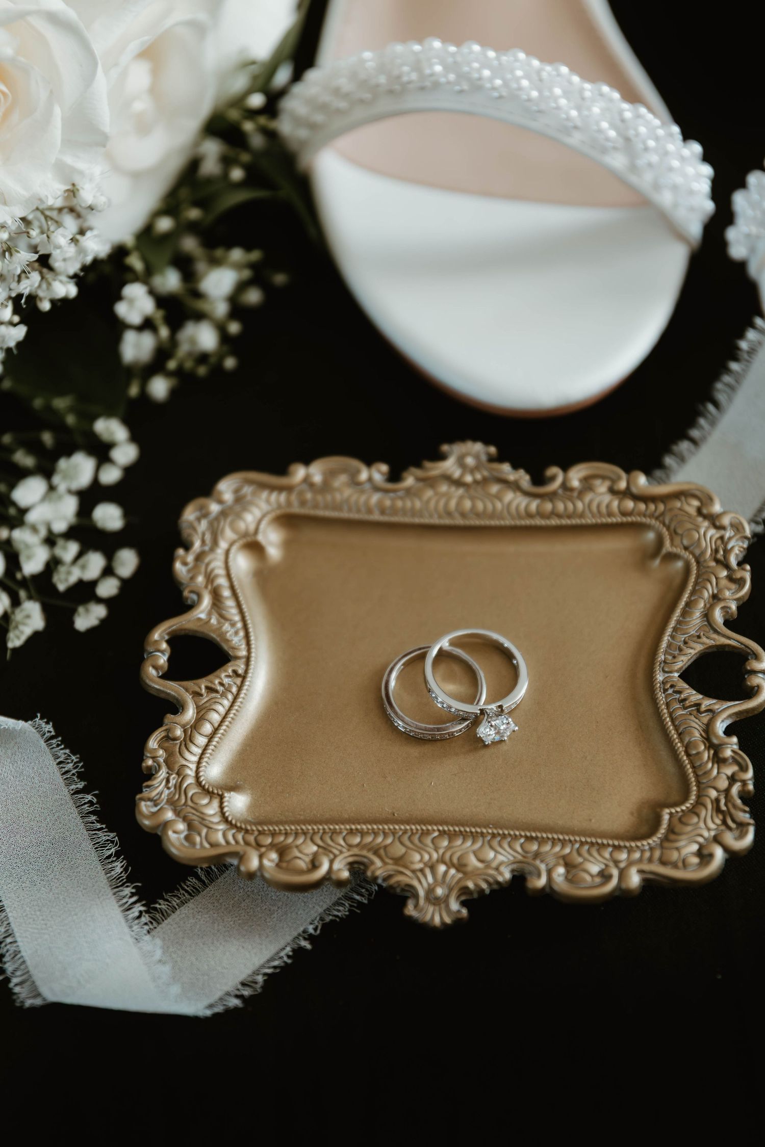 Wedding rings displayed on an ornate gold vintage tray with white flowers and bridal shoe details.