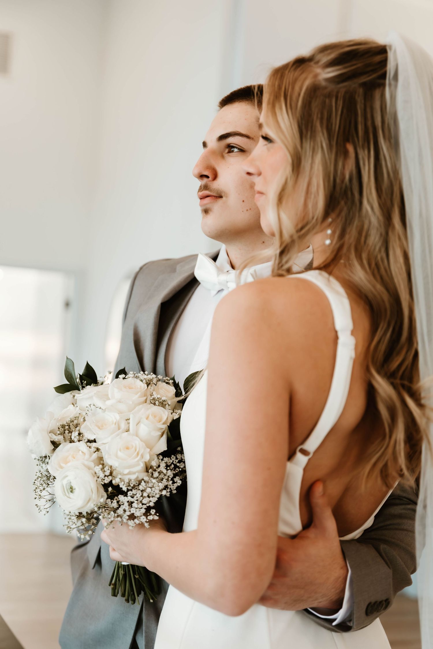 Intimate wedding couple embraces while holding a delicate white rose and baby's breath bouquet.