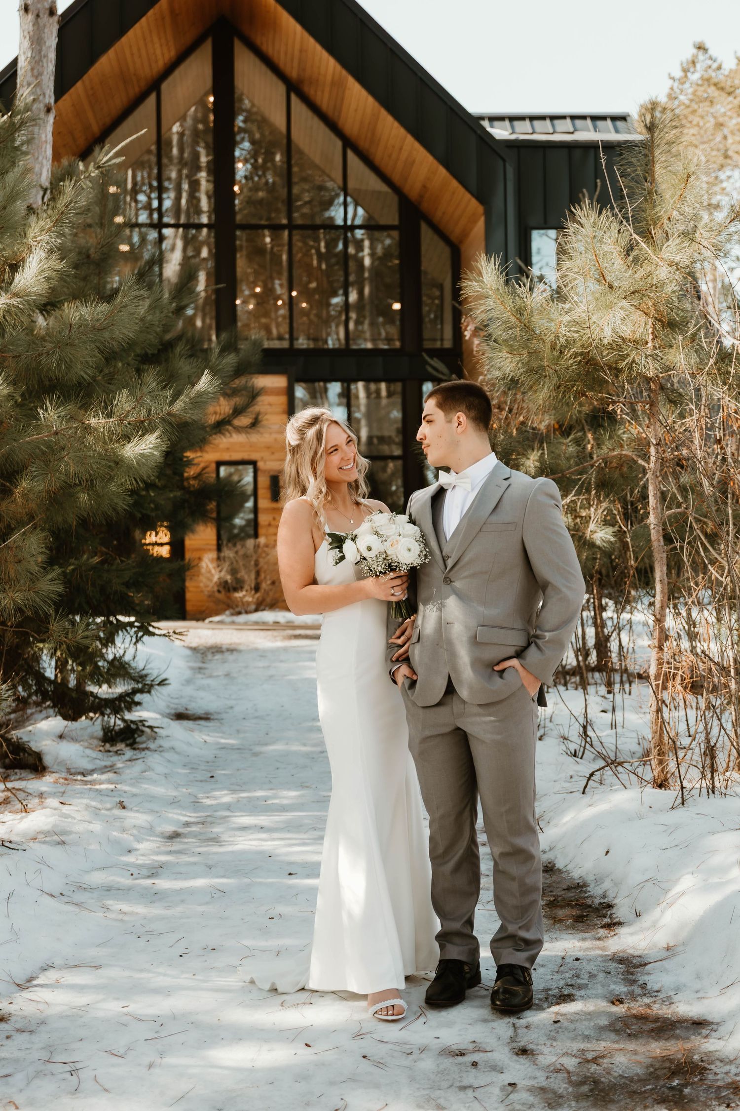 A winter wedding photo in front of a rustic A-frame cabin surrounded by snow and pine trees.