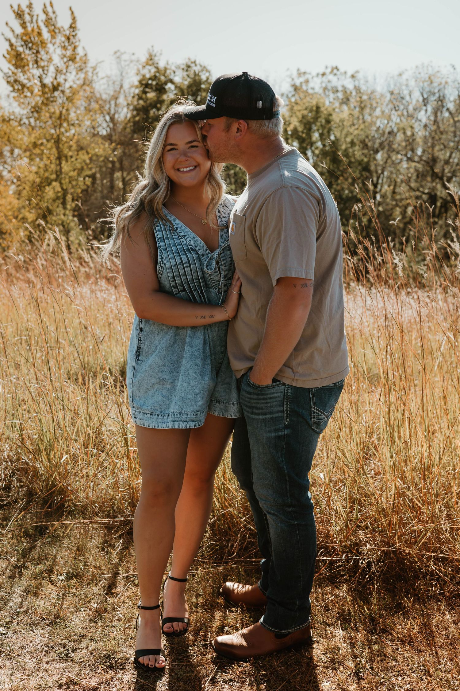 A couple shares an intimate moment together in a sunlit field with tall grass and autumn trees in the background.
