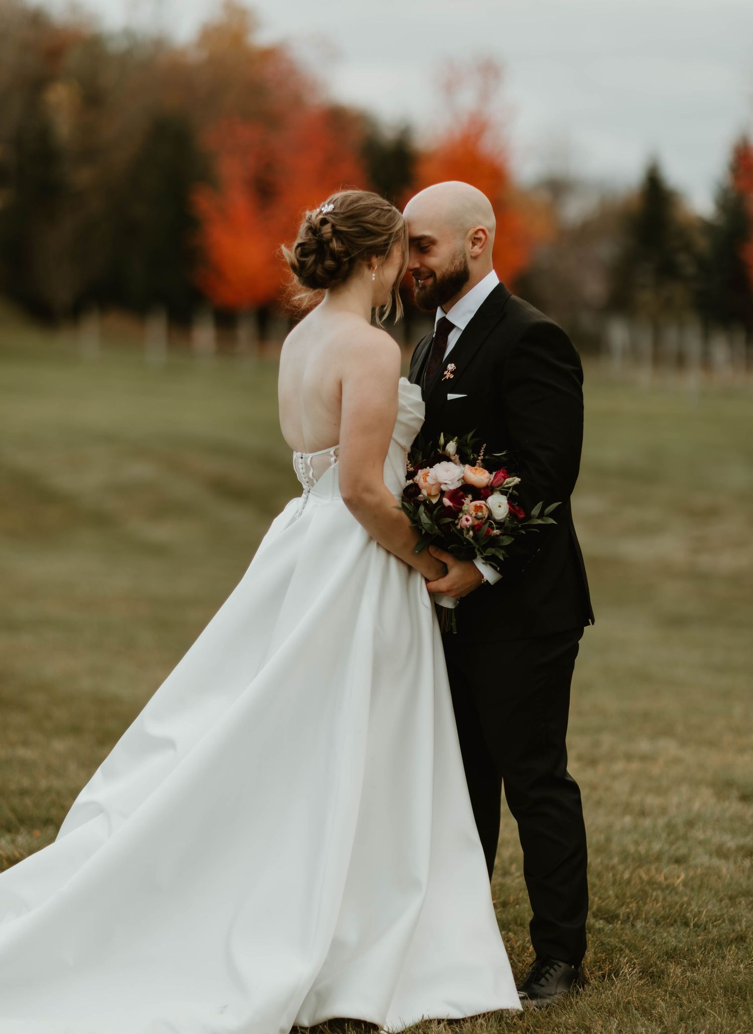 Romantic wedding couple poses in a field during autumn with colorful trees in the distance.