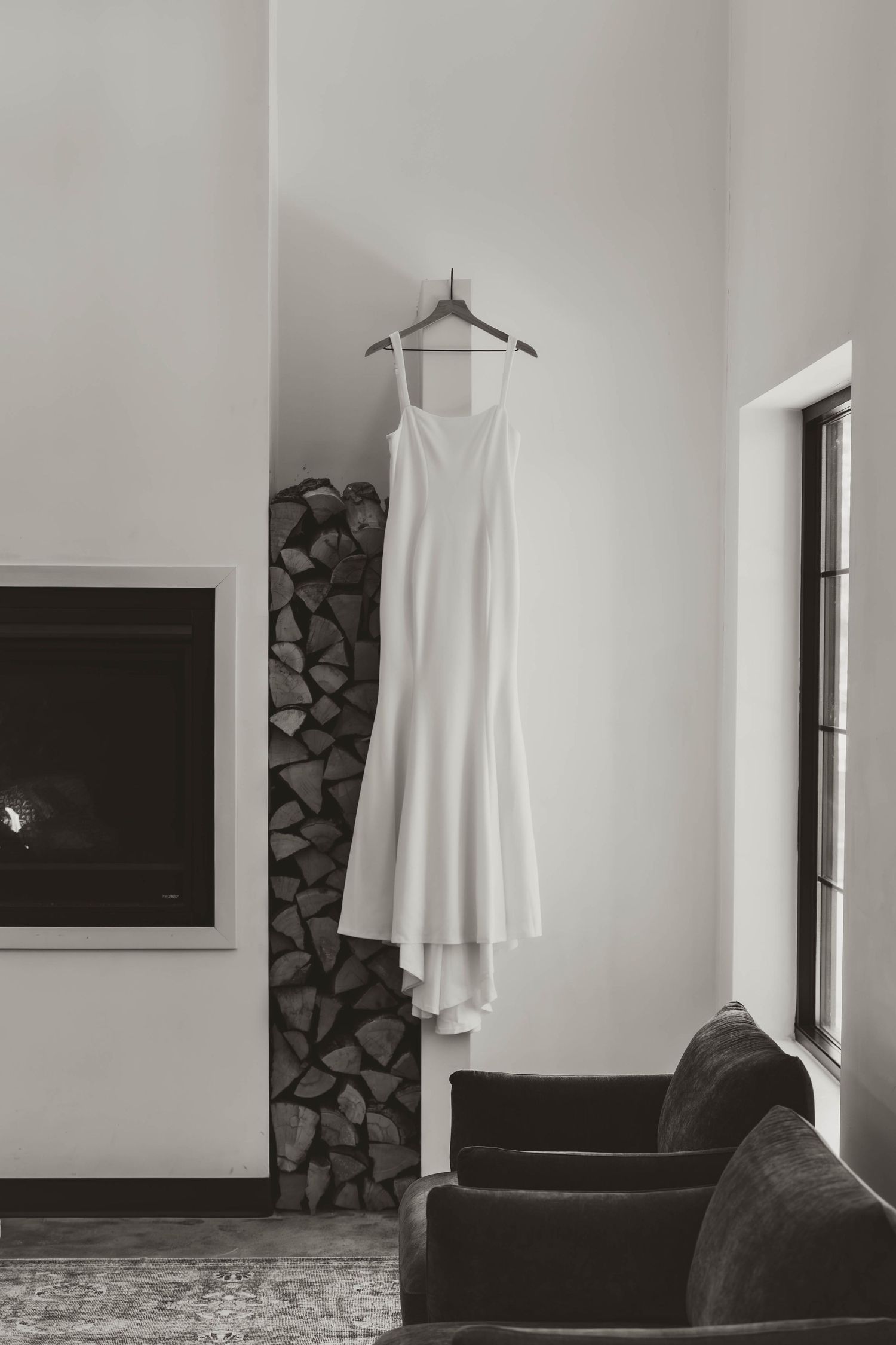 A minimalist white wedding dress hangs on a hanger against a white wall with stacked firewood and dark furniture visible.