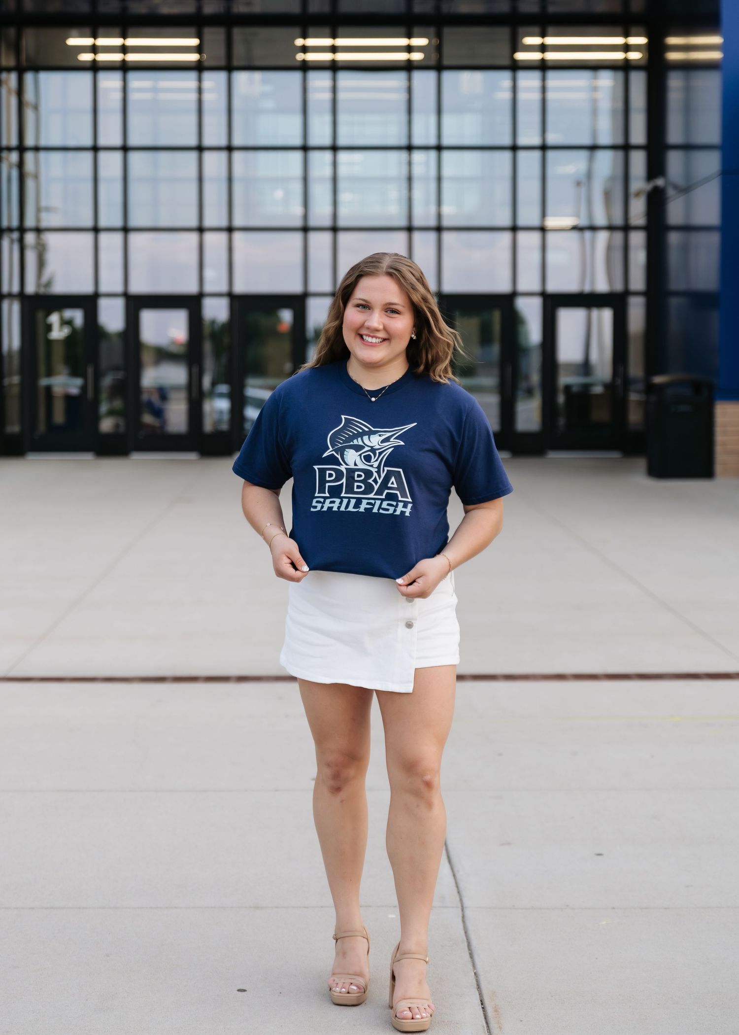 Young woman smiling in a navy PBA Sailfish t-shirt and white skirt, standing outside a modern building.