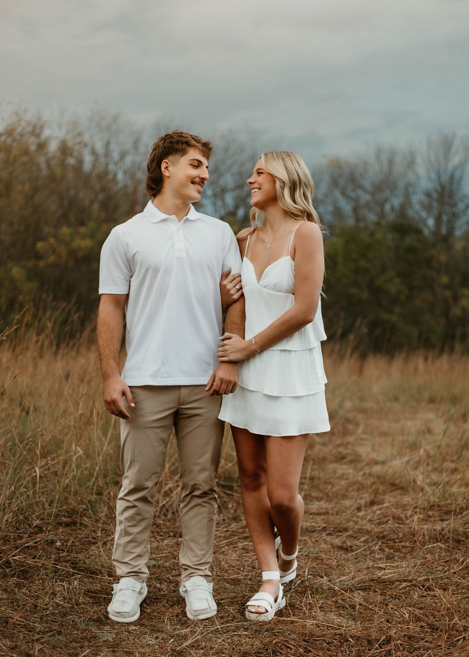 A couple in casual white outfits laughing together in a rustic outdoor field setting at sunset.