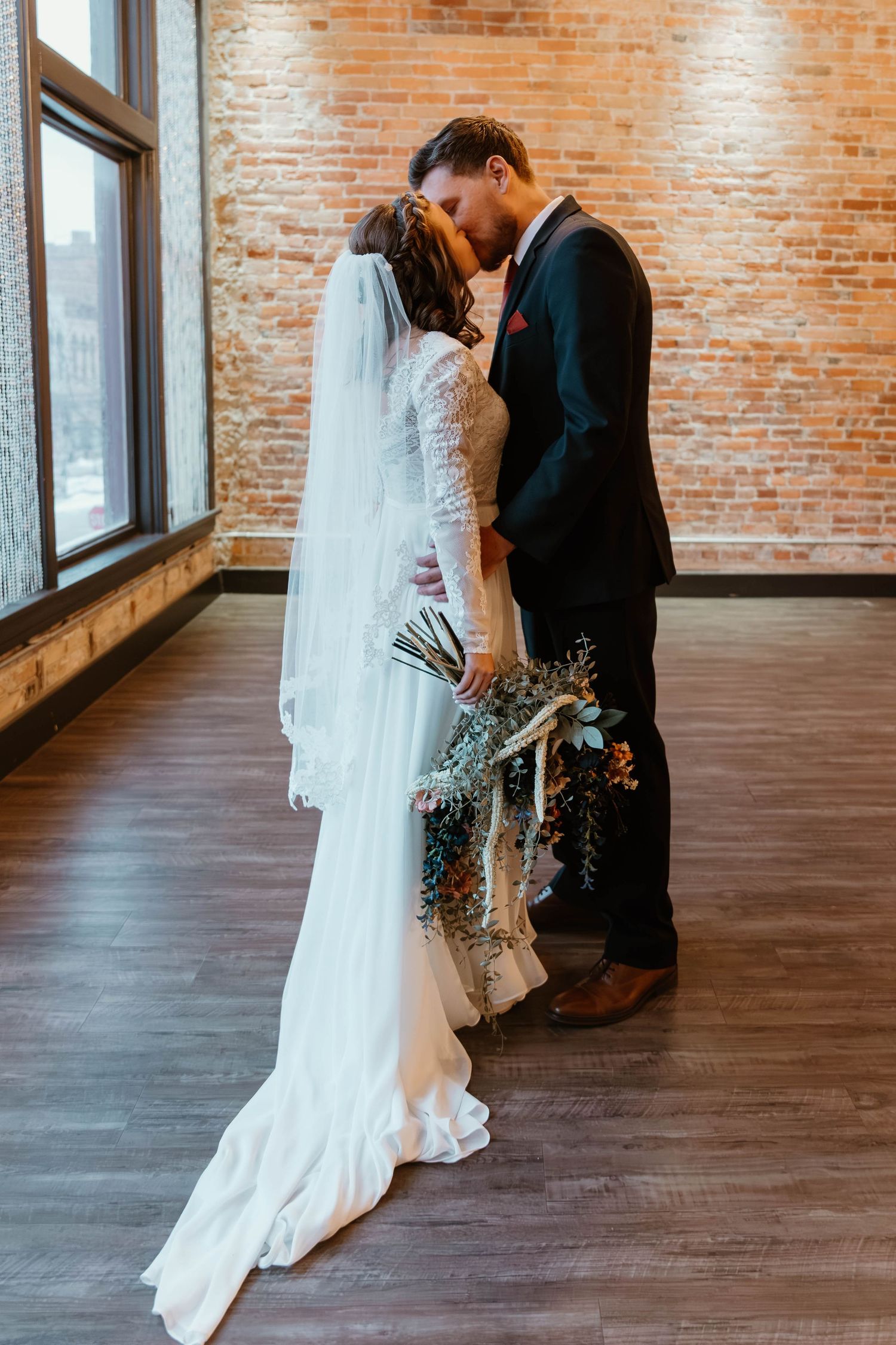 Wedding couple embracing in elegant brick-walled venue with hardwood floors and natural light.