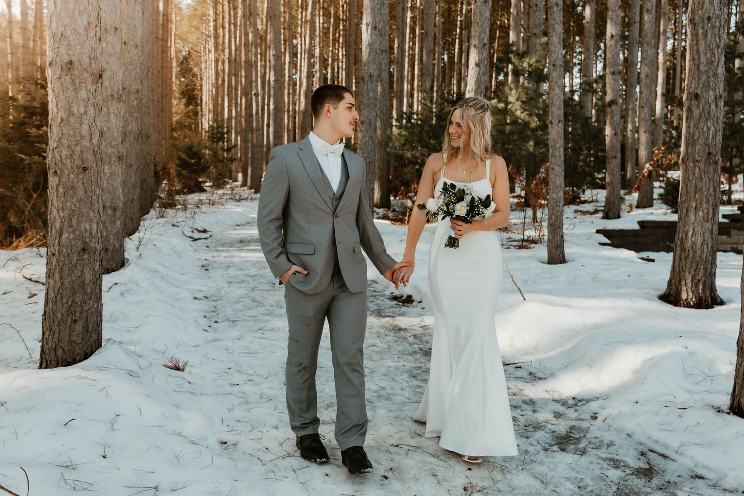 Wedding couple walking through snowy forest path with tall pine trees.