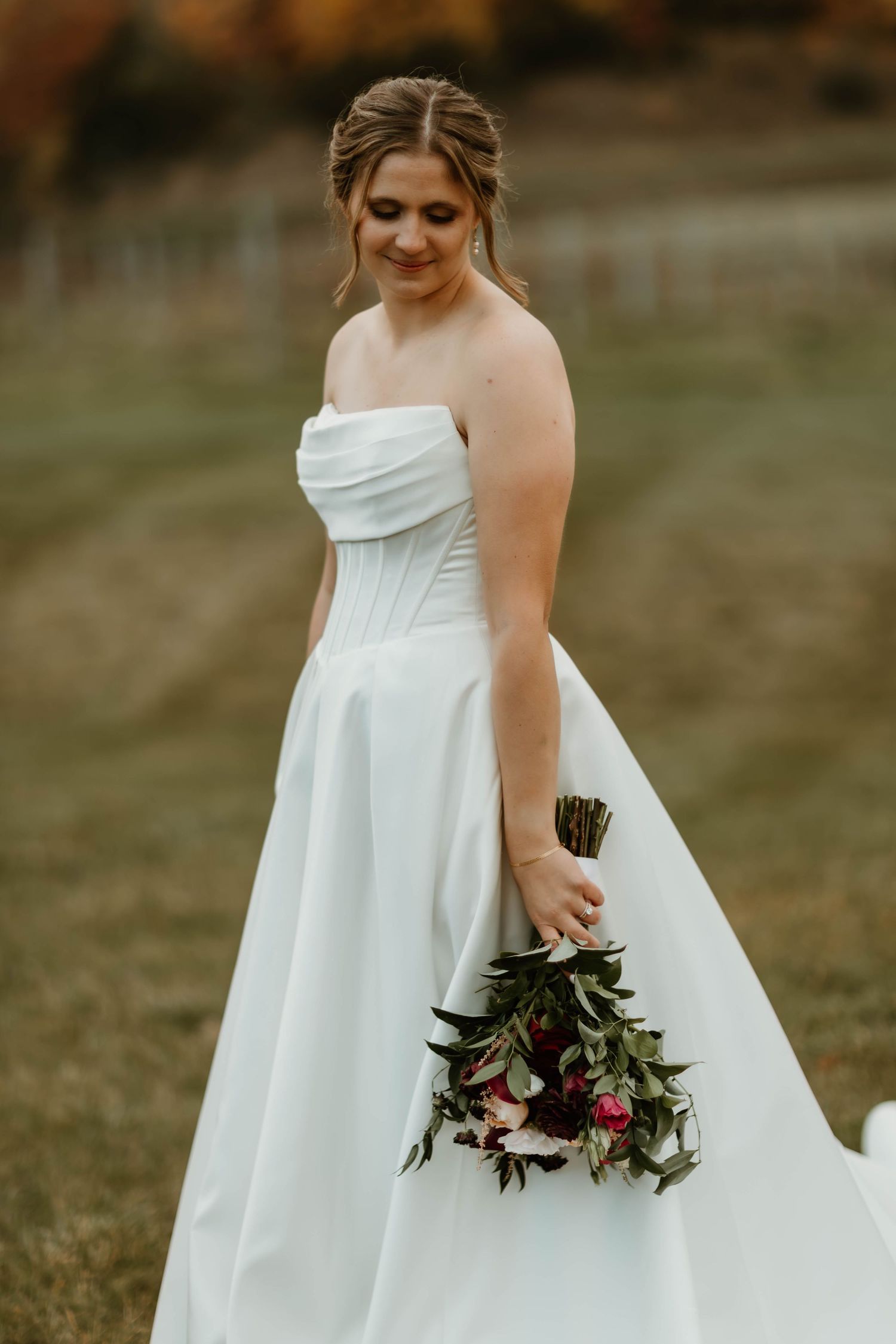 Bride in elegant white strapless wedding gown holds dark floral bouquet with pink accents in outdoor setting.