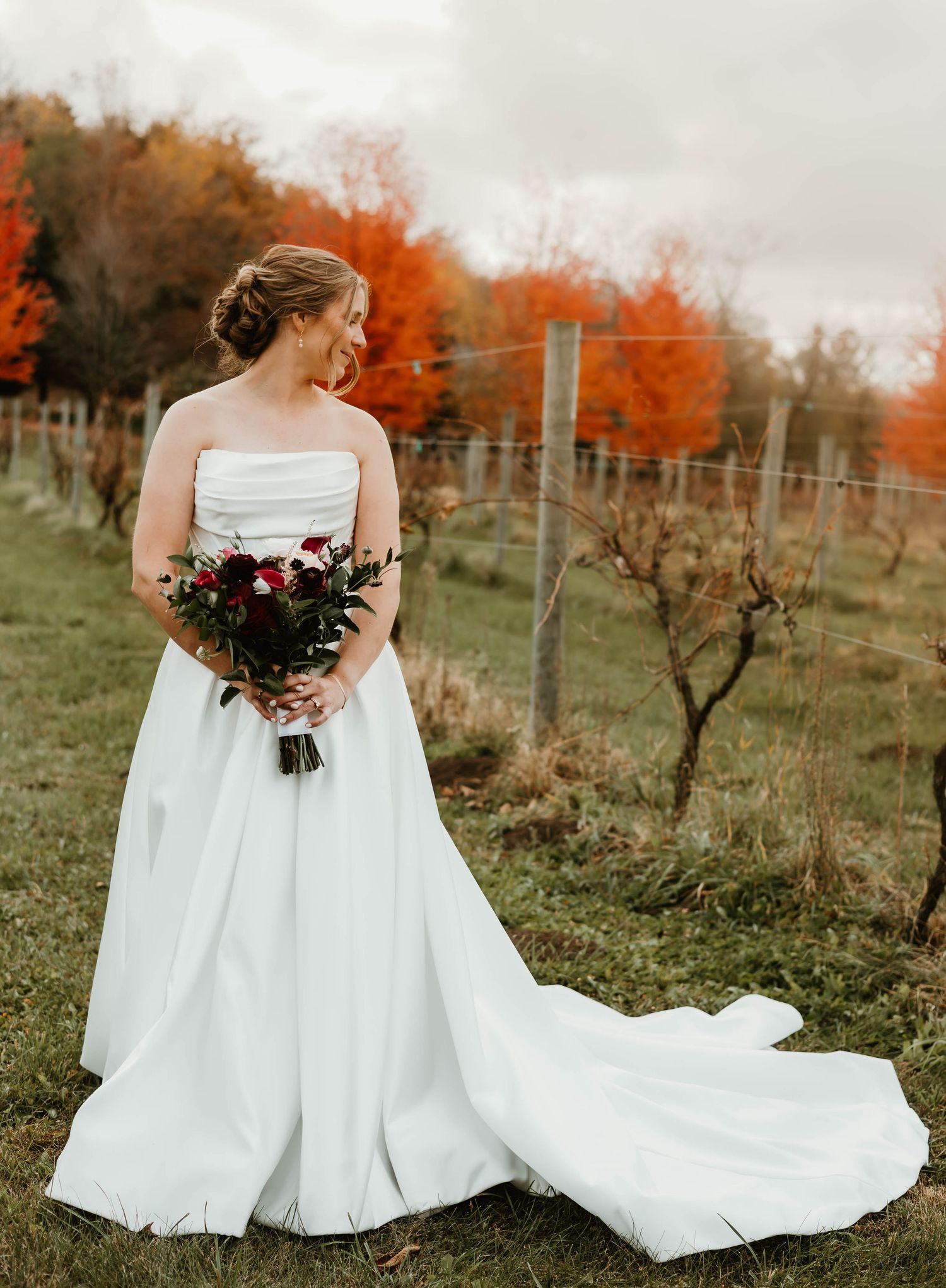 A bride in a white dress with a flowing train walks through a vineyard in fall colors.