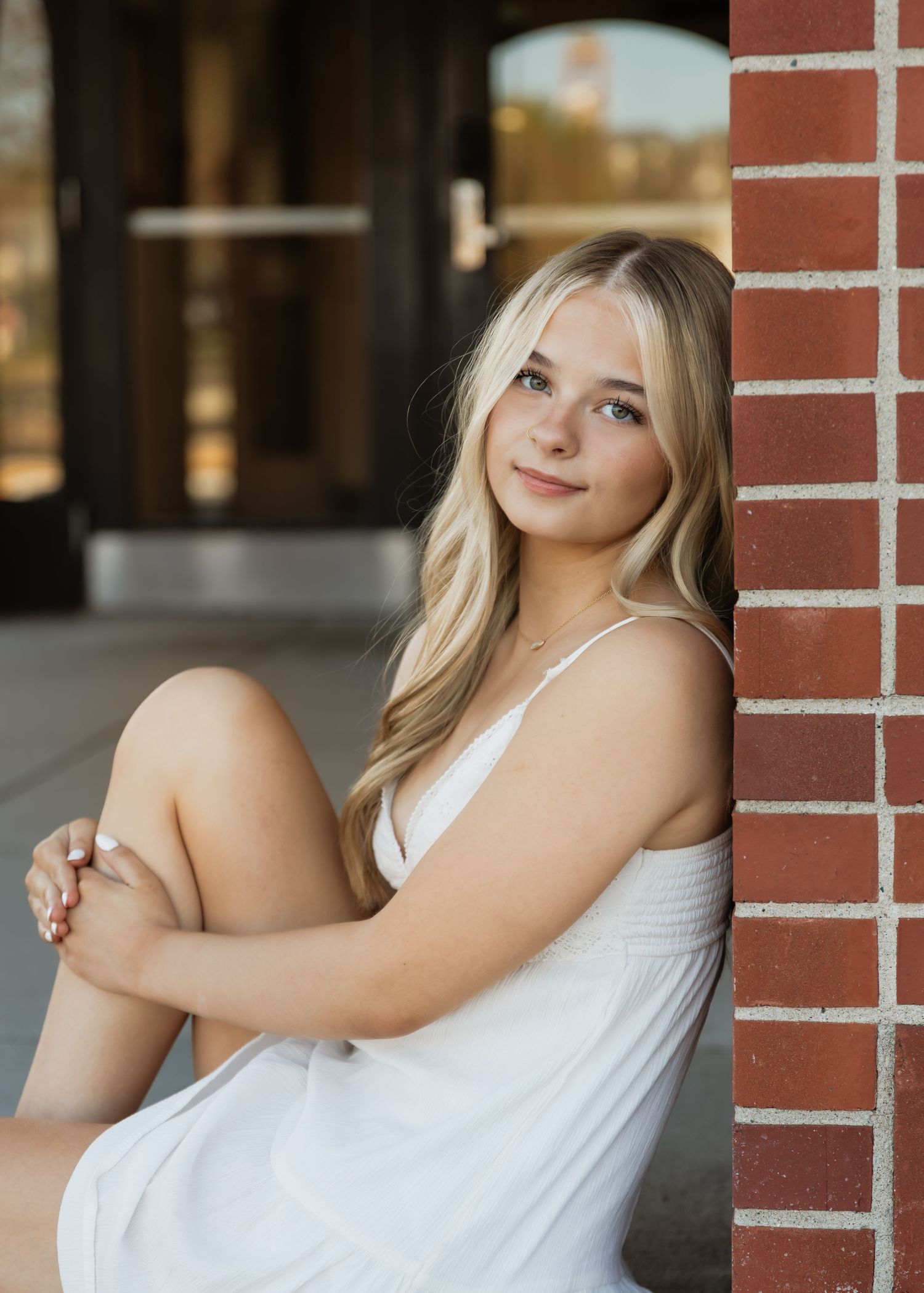 Young blonde woman in white dress poses gracefully against brick column for senior portrait photo.