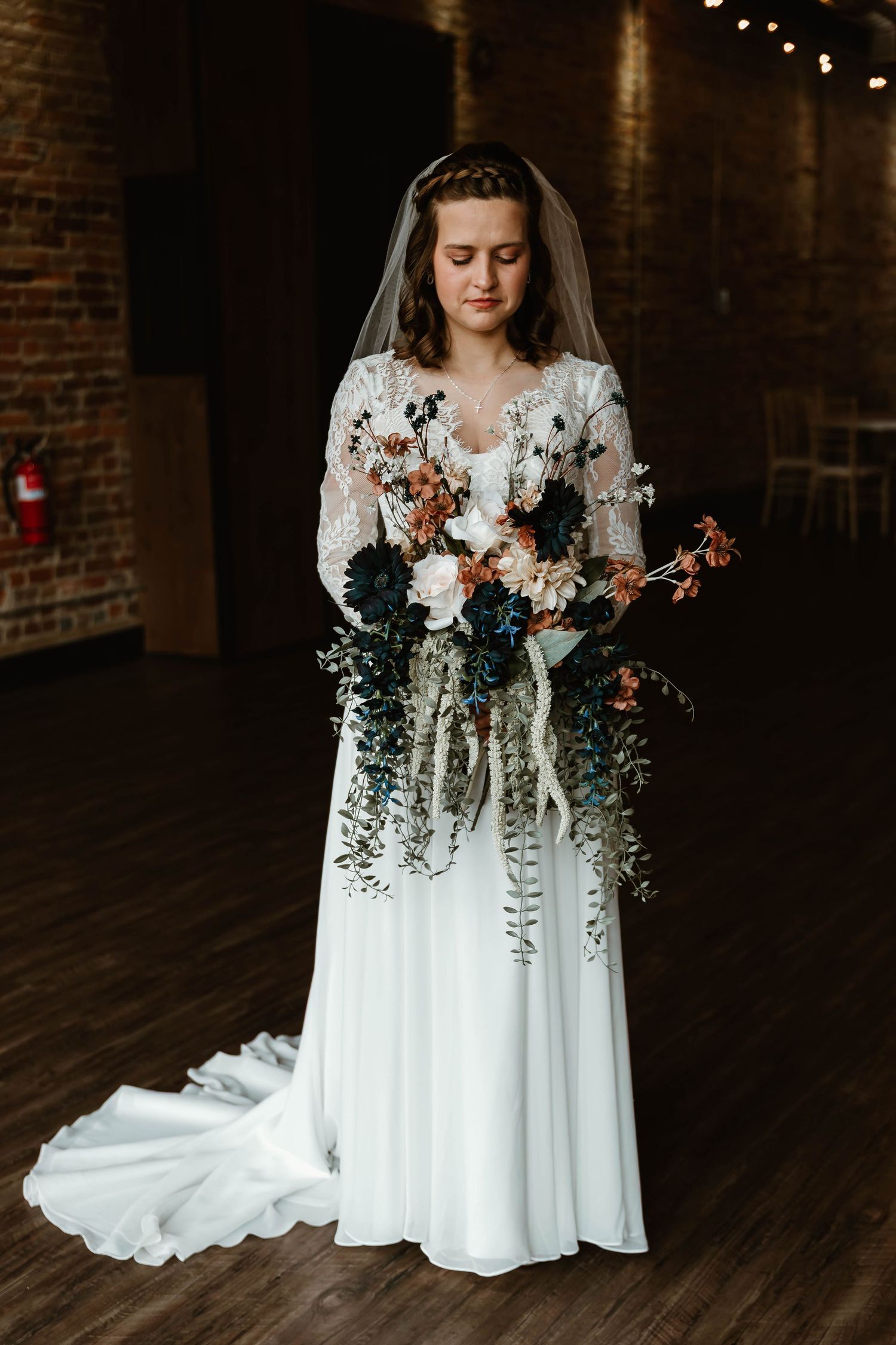 A bride in a flowing white wedding dress holds a romantic bouquet of white and pink flowers against a dark brick wall.