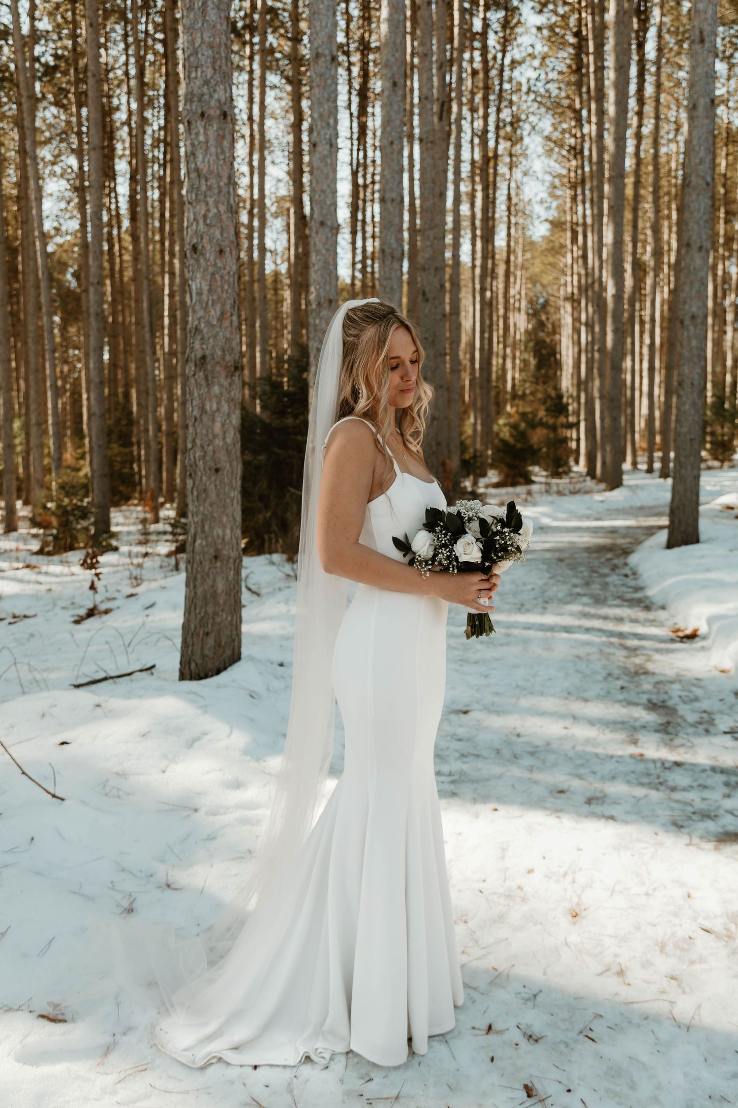 White wedding dress flowing in snowy winter forest with tall pine trees.