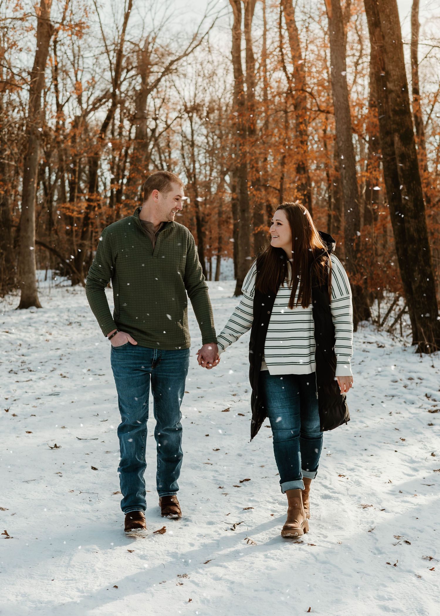 A couple walks hand in hand through a snowy winter forest path during golden hour.