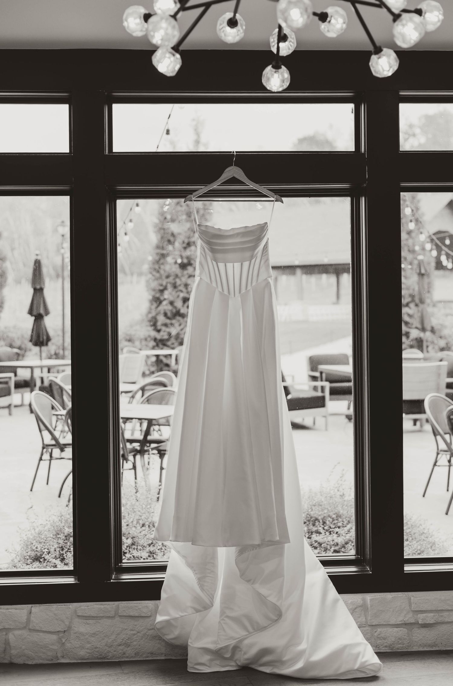 A white wedding dress hangs elegantly in front of large windows with natural light.