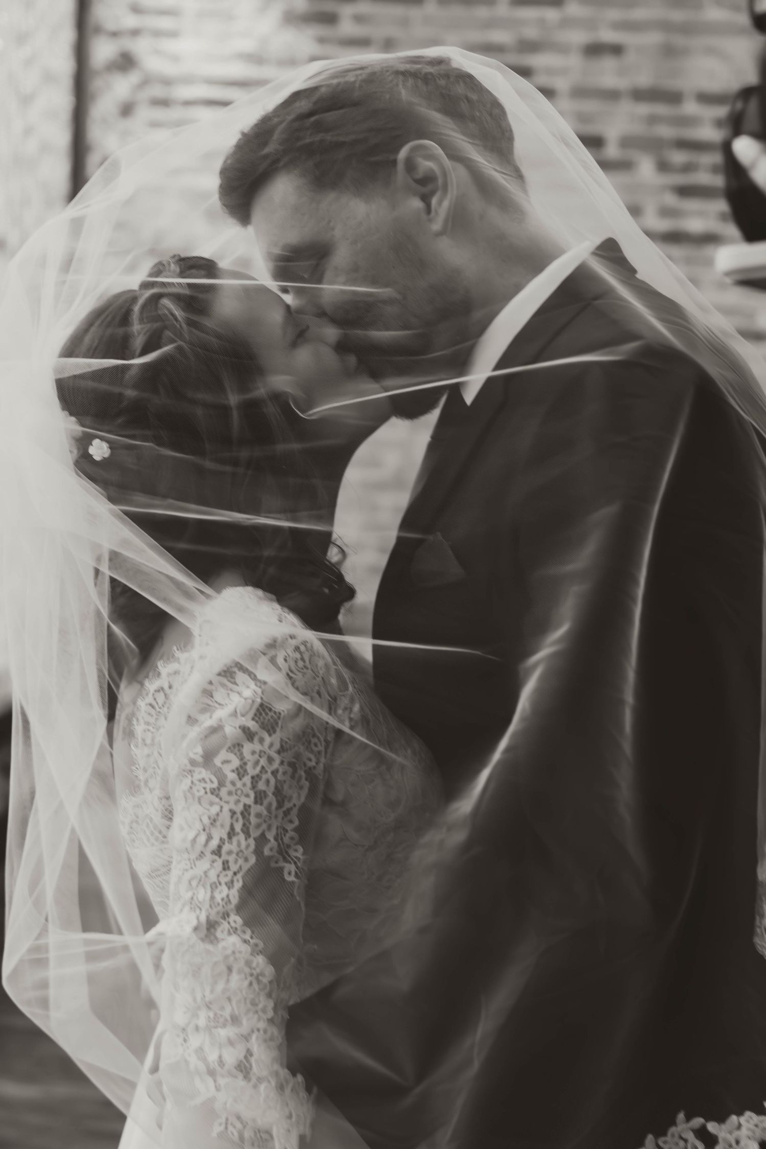 Close-up romantic wedding moment with bride in lace dress and groom in dark suit against brick backdrop.
