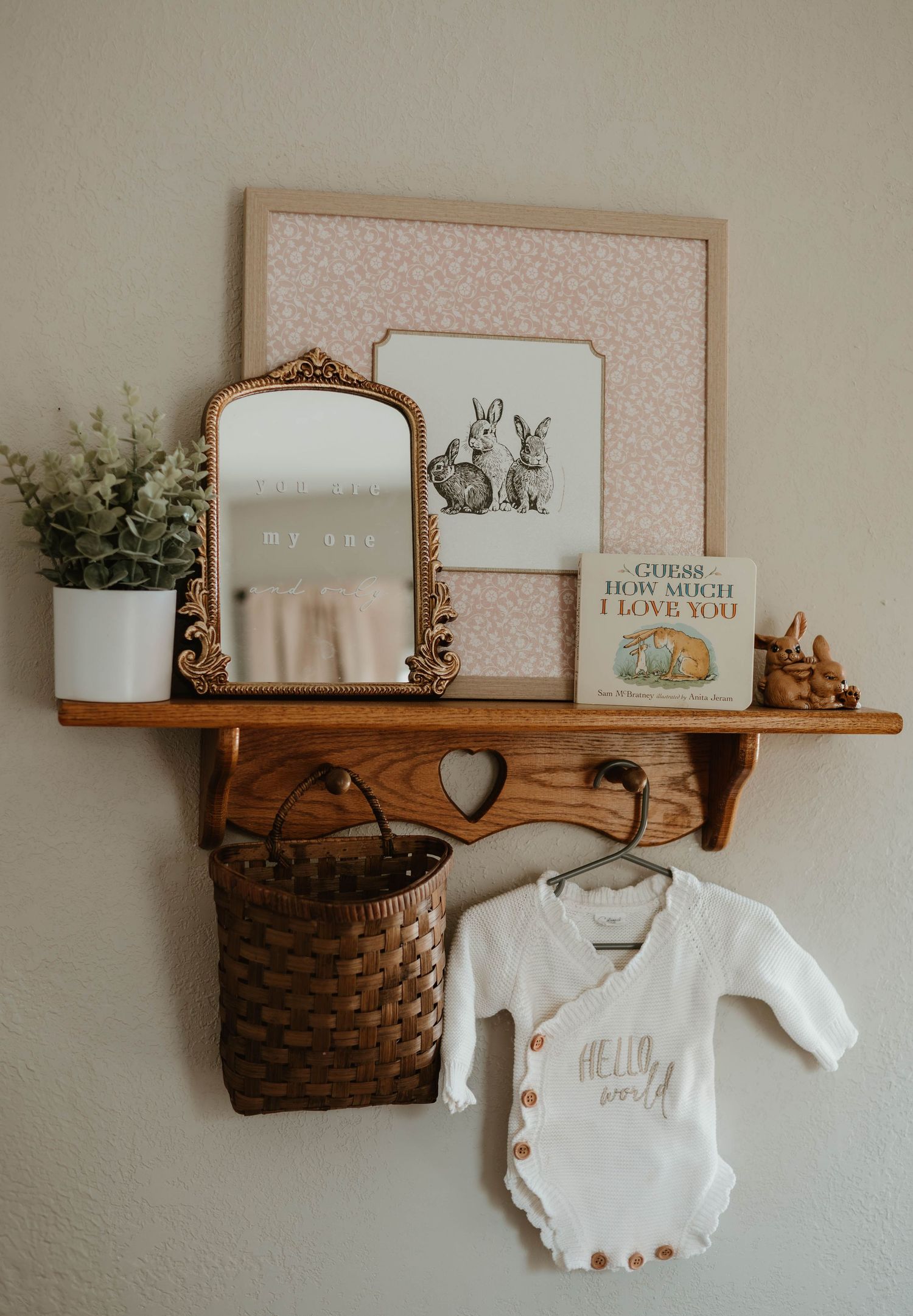 Rustic wooden nursery shelf with bunny art, gold mirror, basket, and Hello World baby onesie for a charming baby girl room.