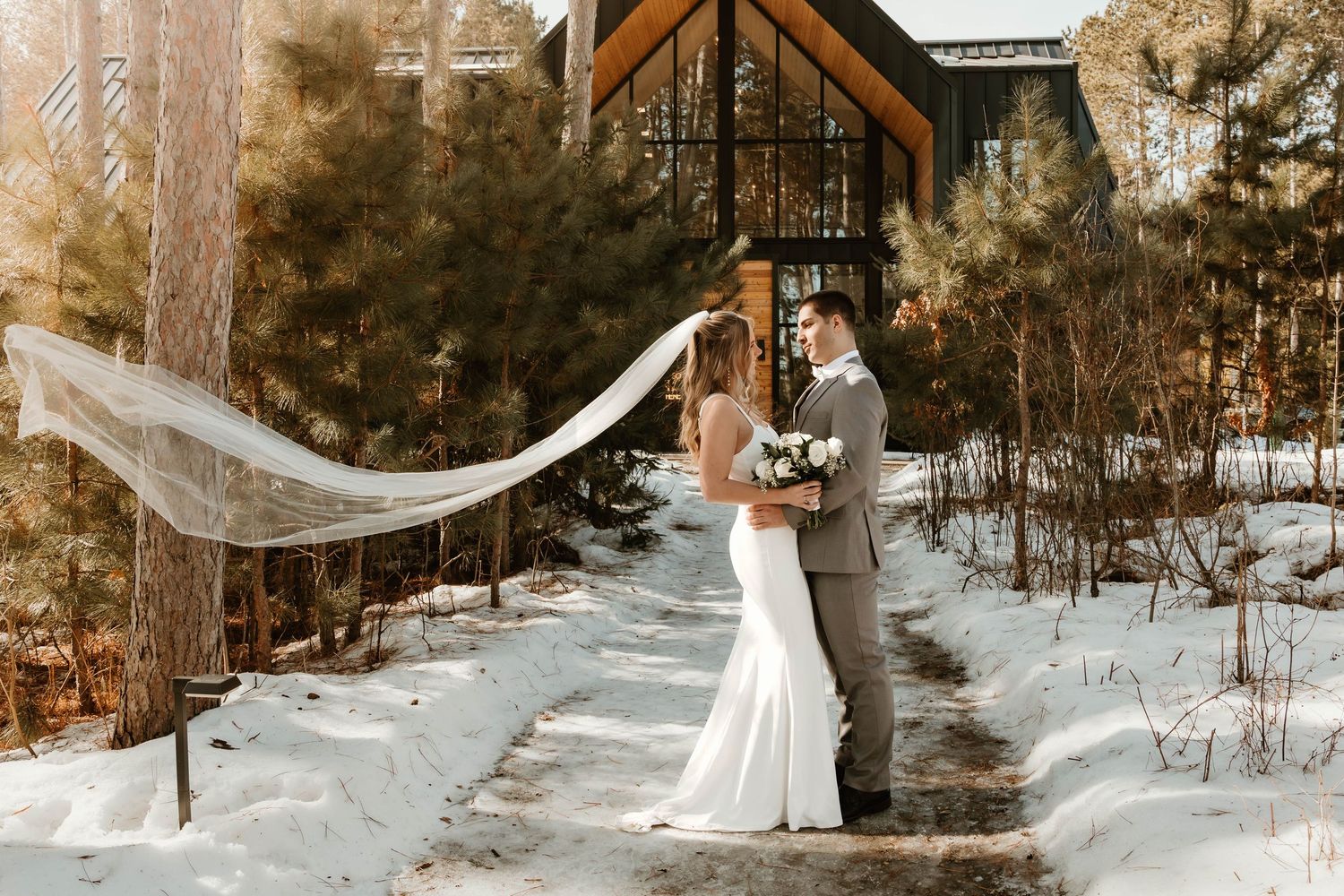 Bride and groom stand in snowy forest path, her veil flowing in wind, with rustic chapel backdrop in winter.
