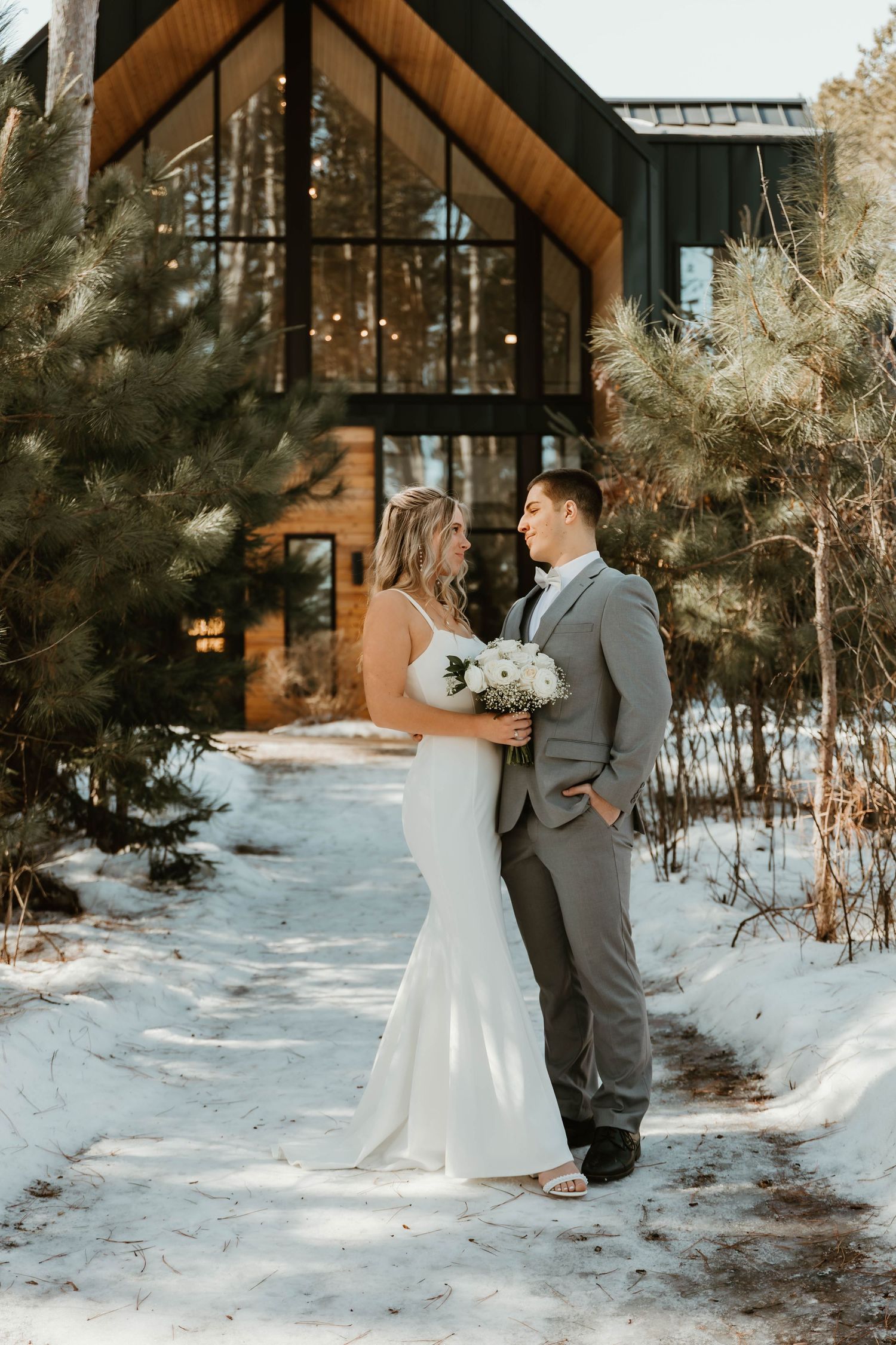 Winter wedding portrait in front of modern wooden lodge with snow-covered ground.