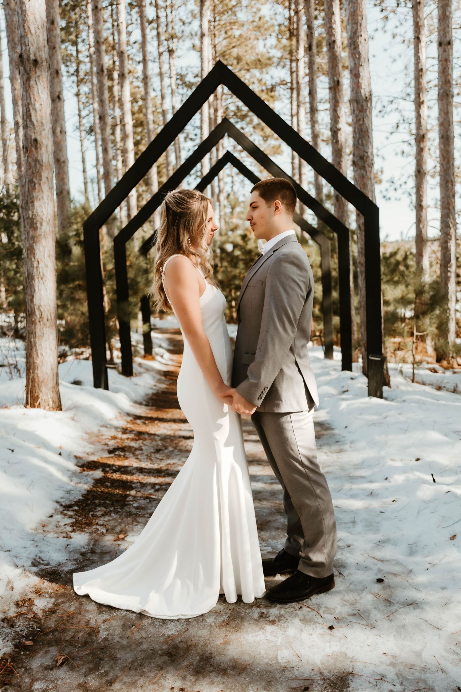 Wedding couple standing under geometric black arches in snowy forest setting.