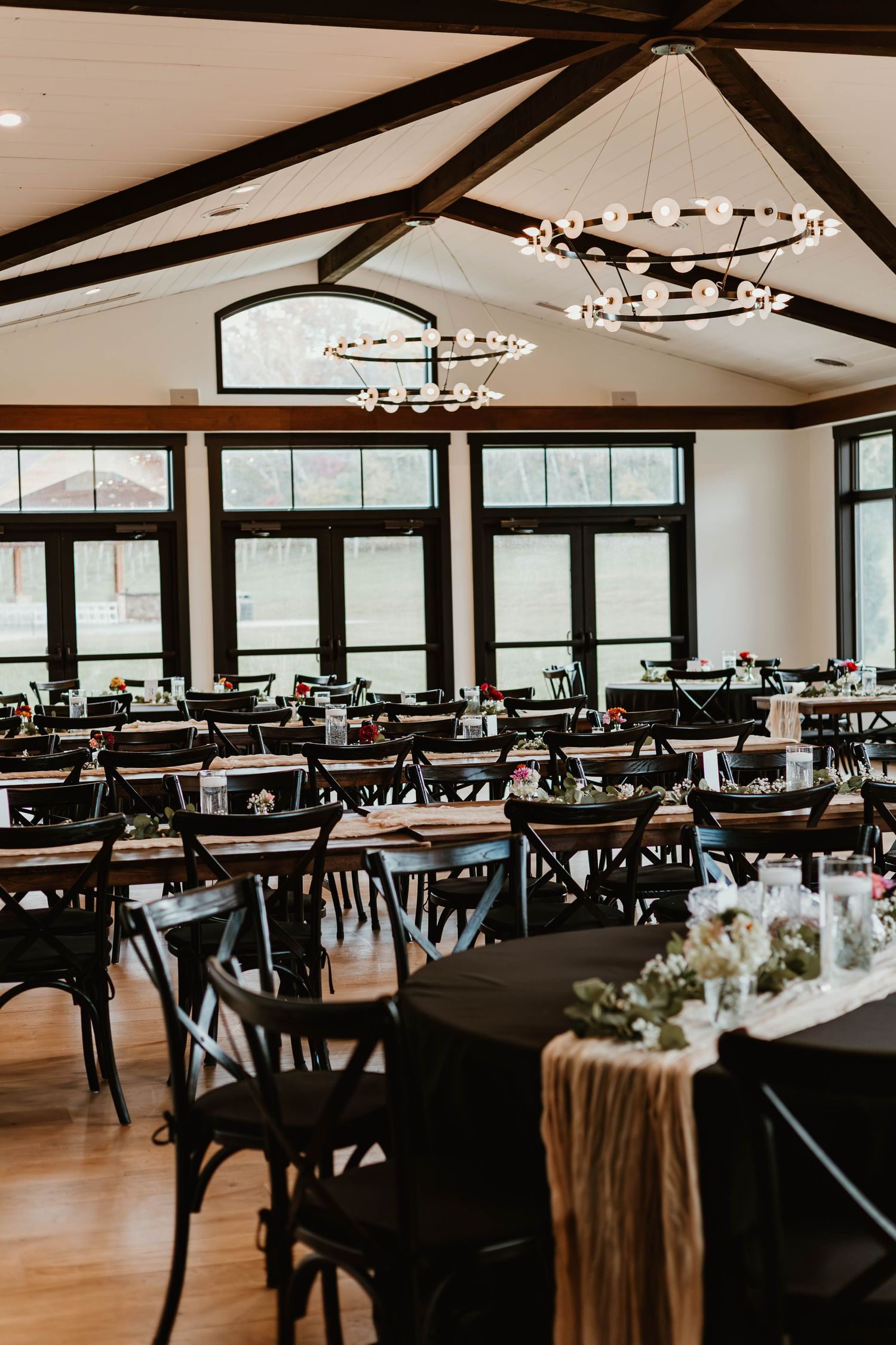 Modern wedding reception hall with exposed wooden beams, black chairs, and elegant chandeliers set for a formal event.