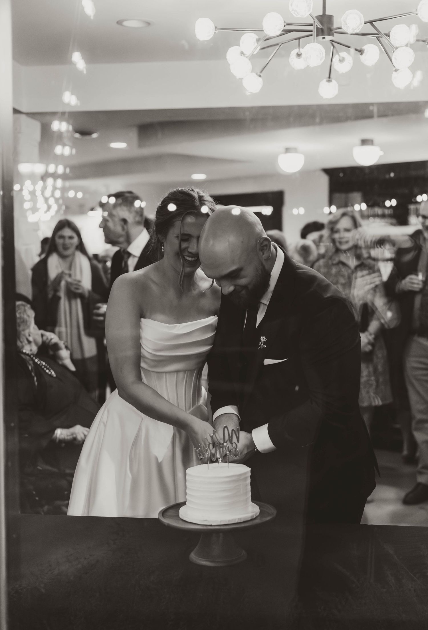 Wedding reception scene with guests gathered around couple cutting cake.