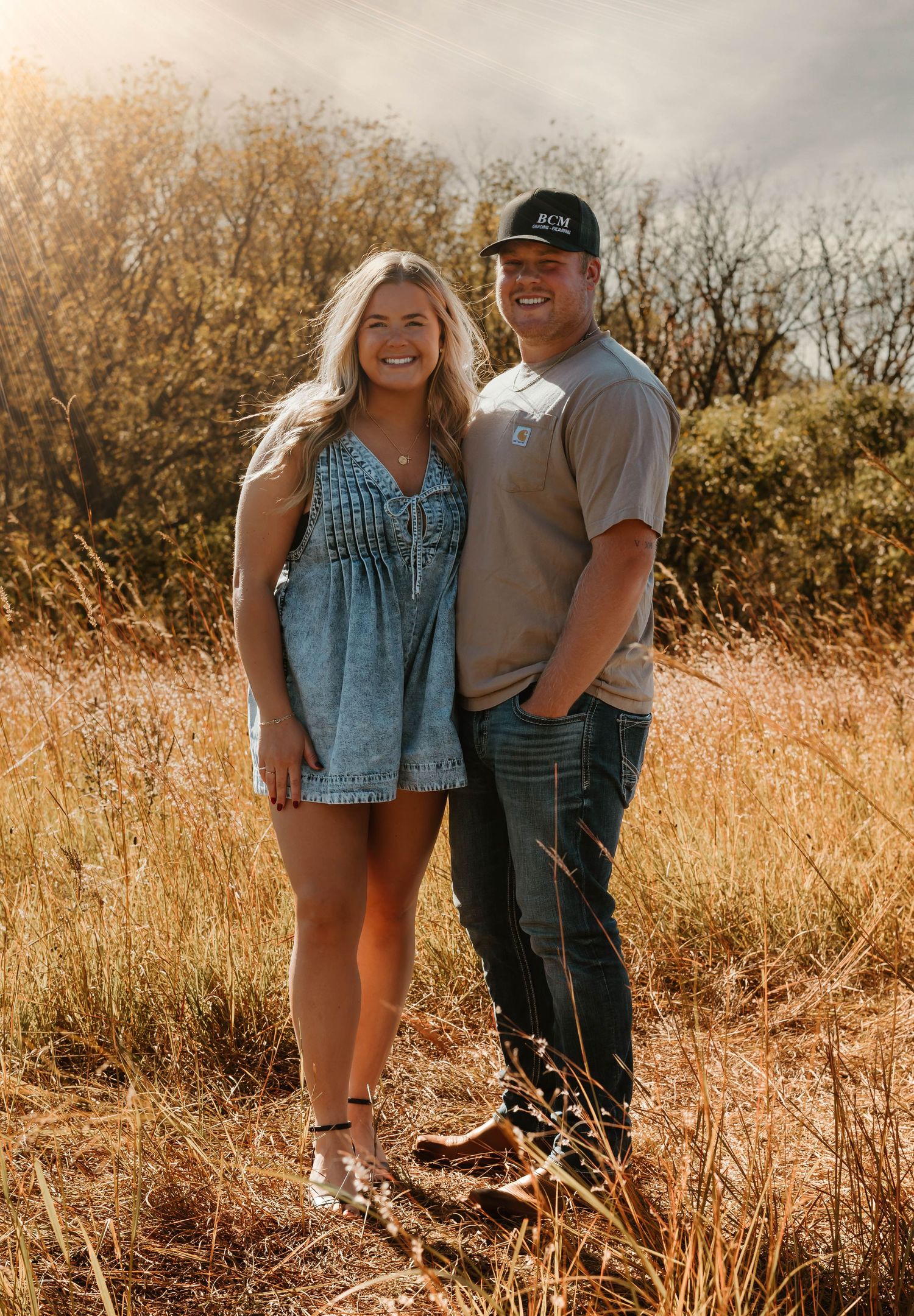 A couple poses together in a rustic outdoor setting with tall golden grass and autumn foliage.