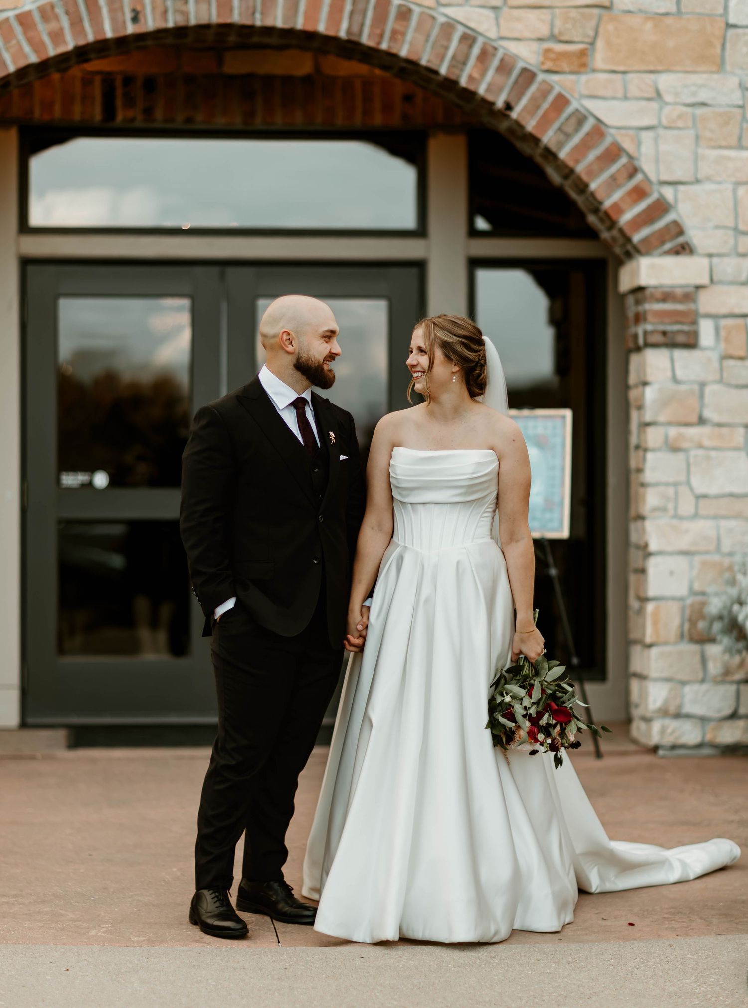 Wedding couple stands together outside stone building, bride in white gown holding burgundy bouquet.