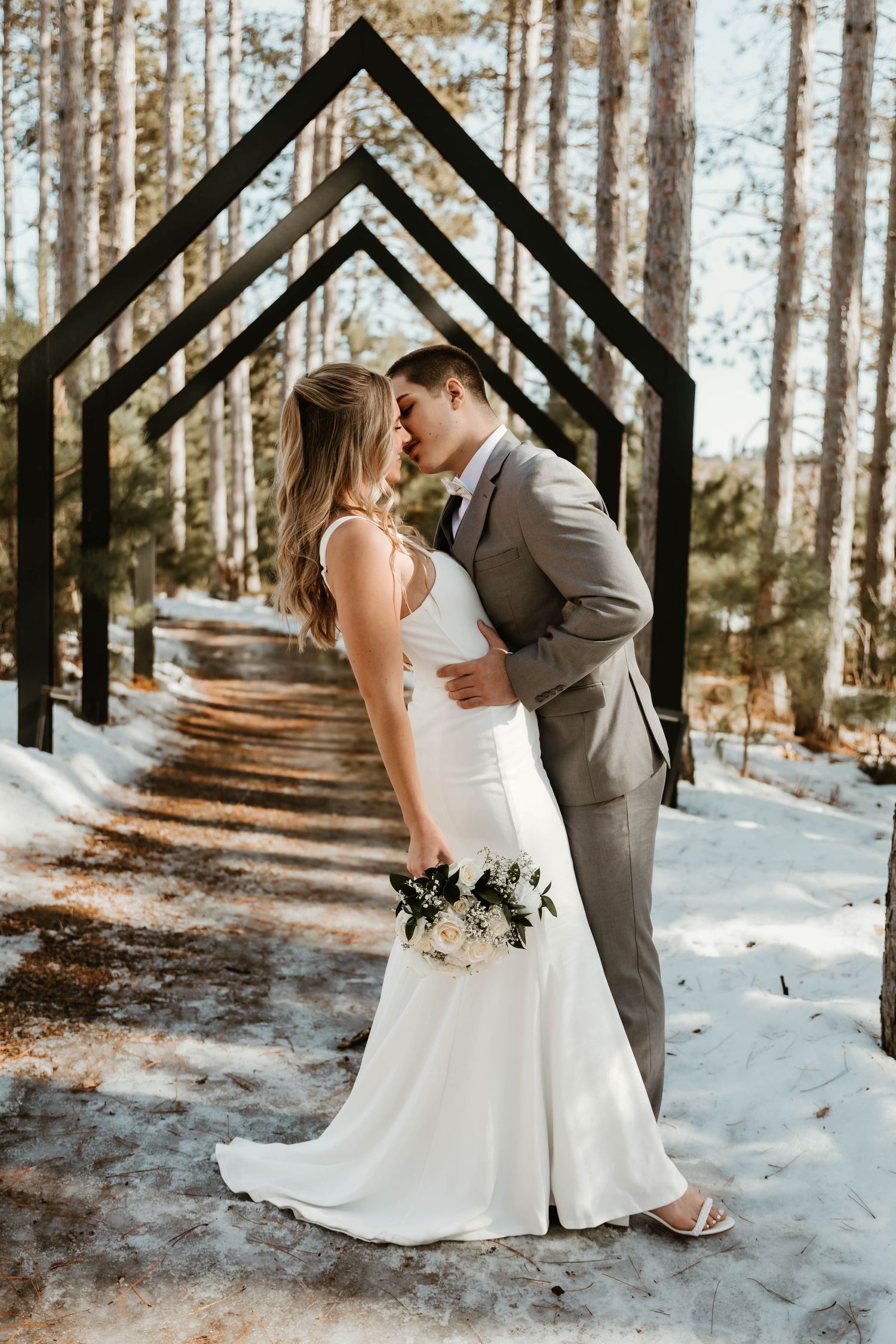 Newlyweds share an intimate moment on a snow-covered forest path lined with tall pine trees.