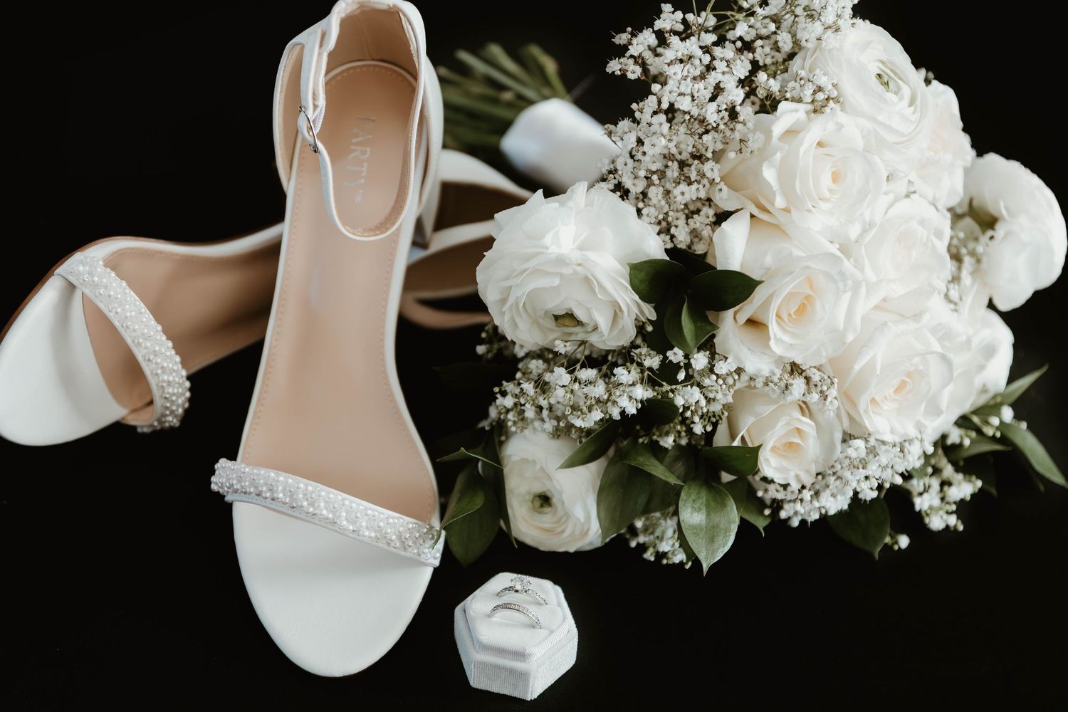 White bridal shoes and floral arrangements displayed with greenery against a black background.