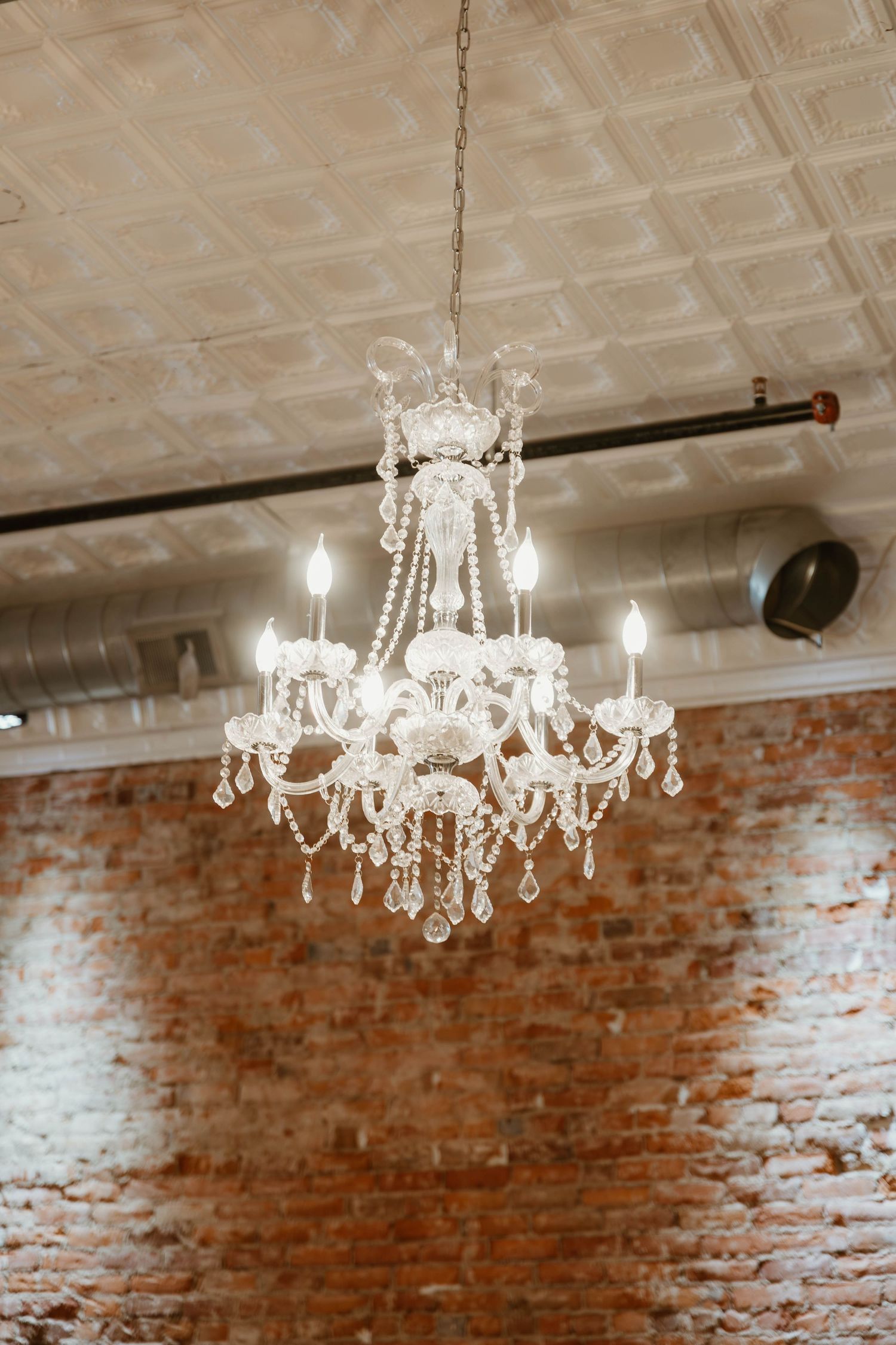Row of crystal chandeliers hanging from a tin ceiling above exposed brick walls in an event space.