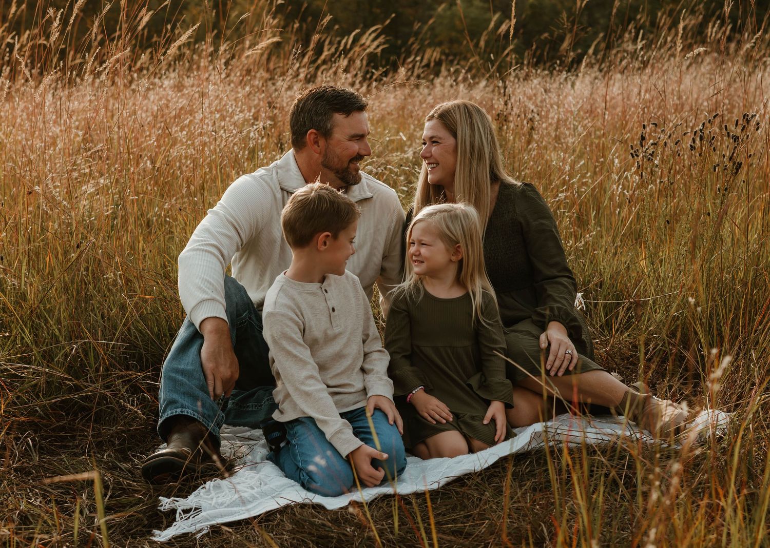 Family of four sitting together on blanket in tall autumn grass.