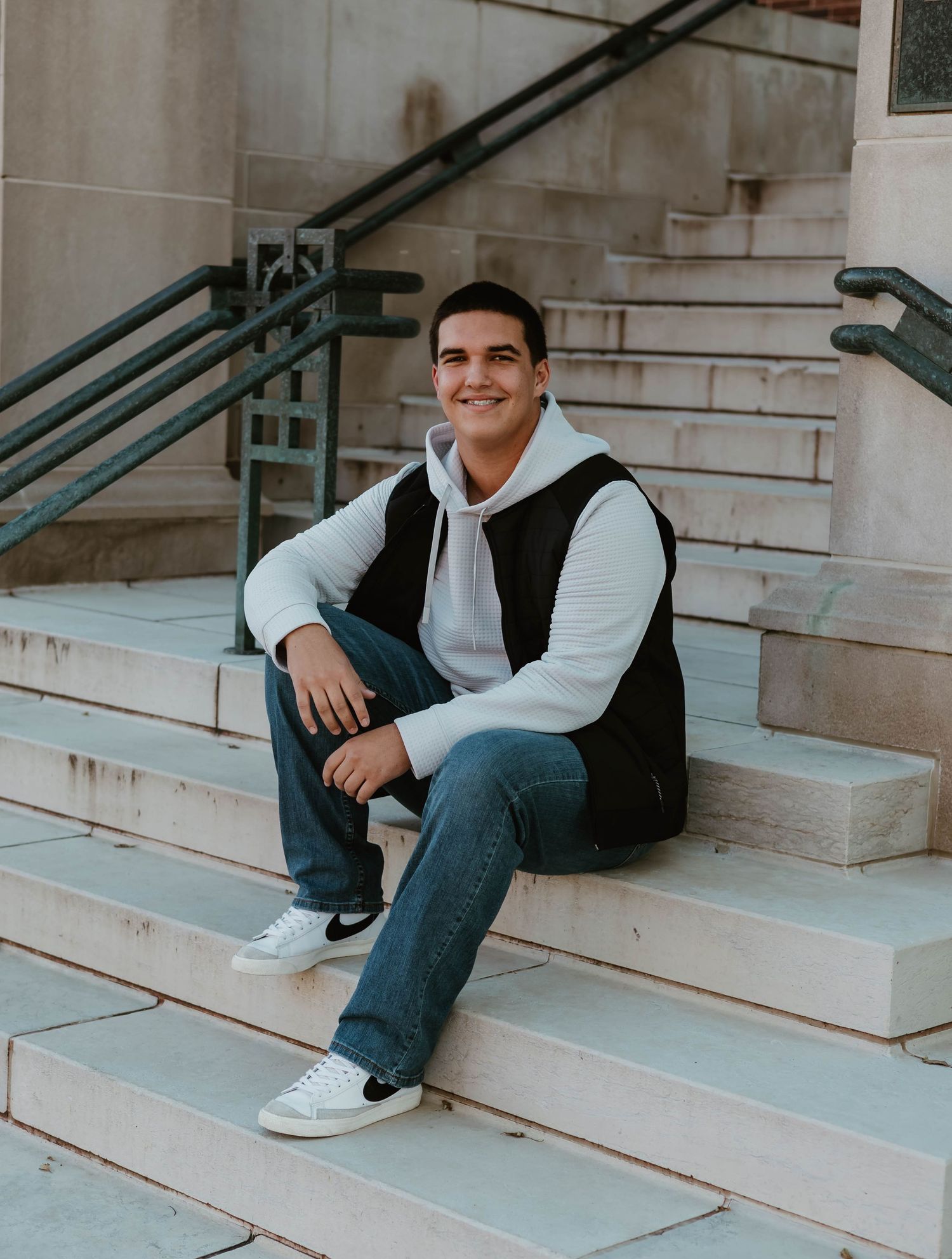 Young man in black vest and white hoodie sitting on stone steps, smiling casually in jeans and white sneakers.