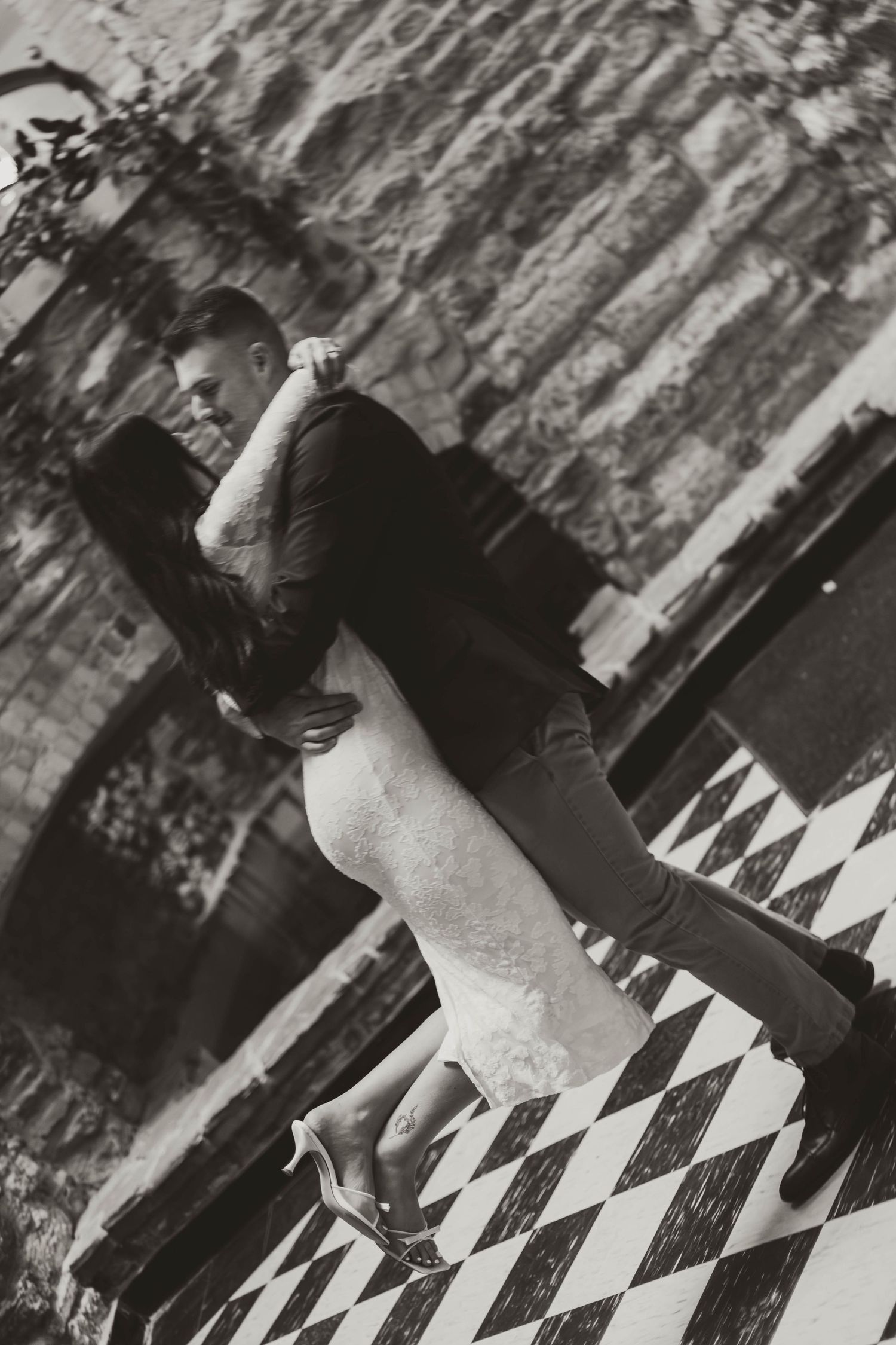 A black and white photo of a couple standing on a geometric patterned floor against a stone wall.
