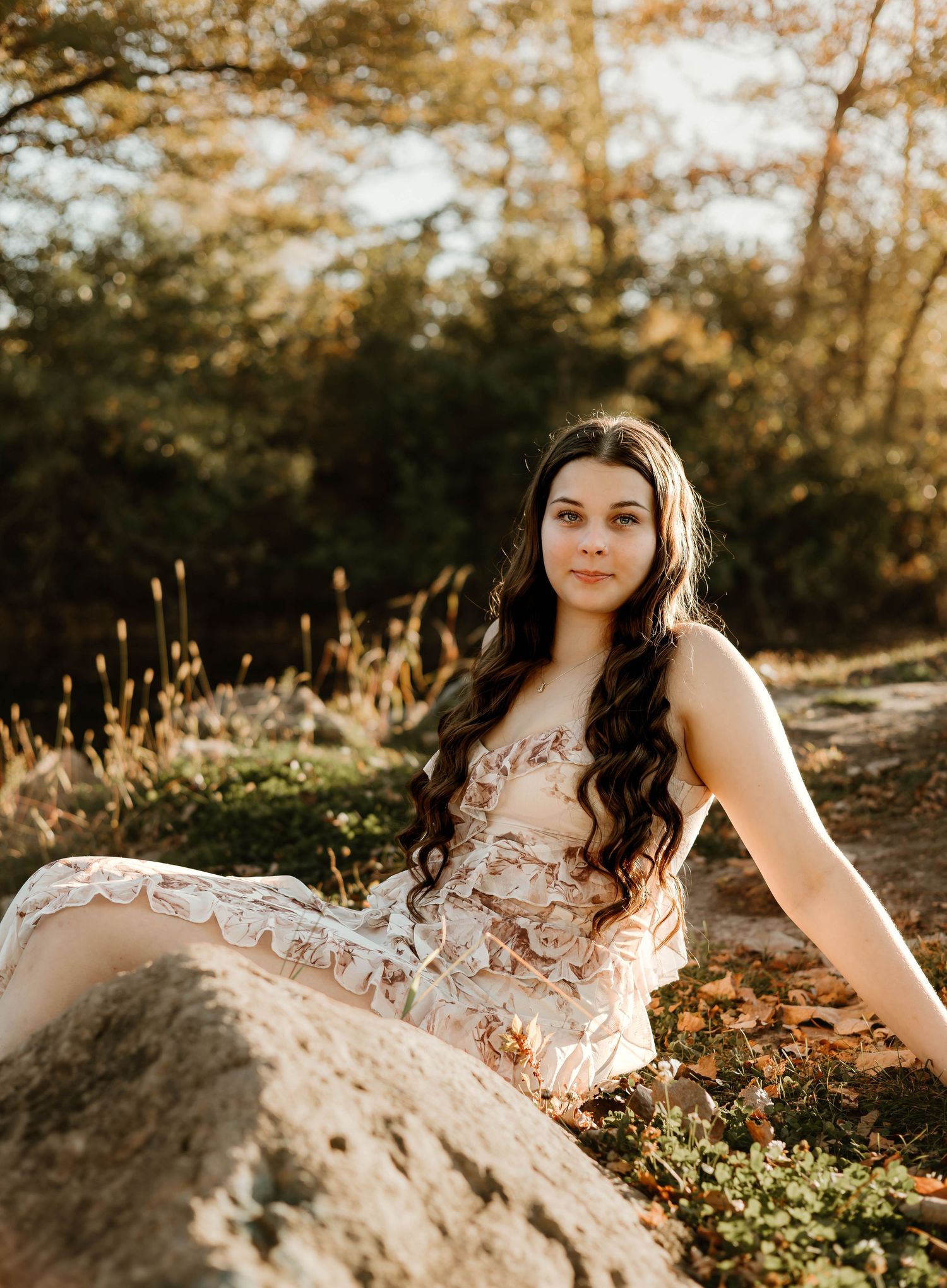Young woman in lace dress sitting on rocks in golden sunlight surrounded by autumn foliage.