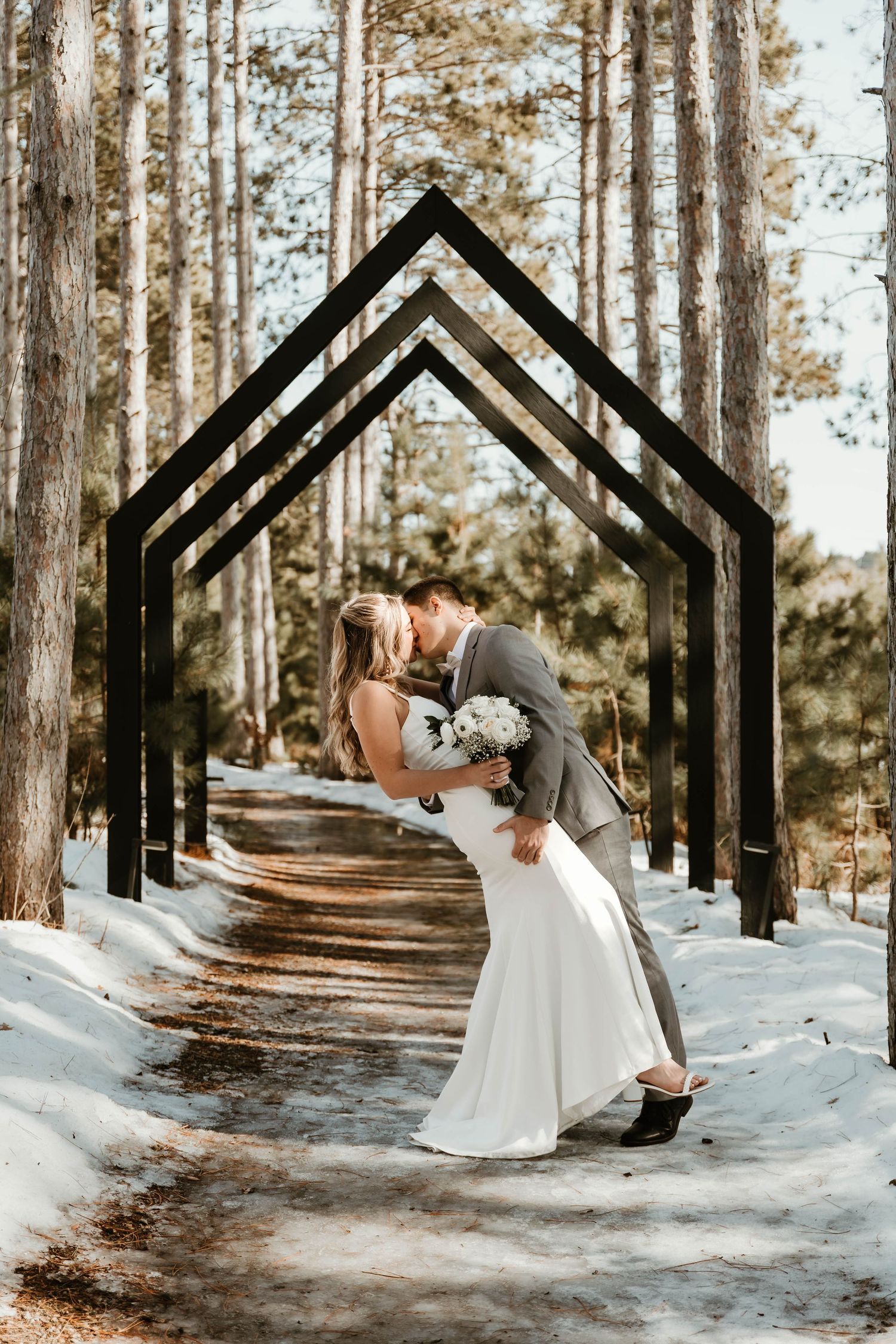 Romantic dip pose under geometric black arches in snowy winter forest setting.