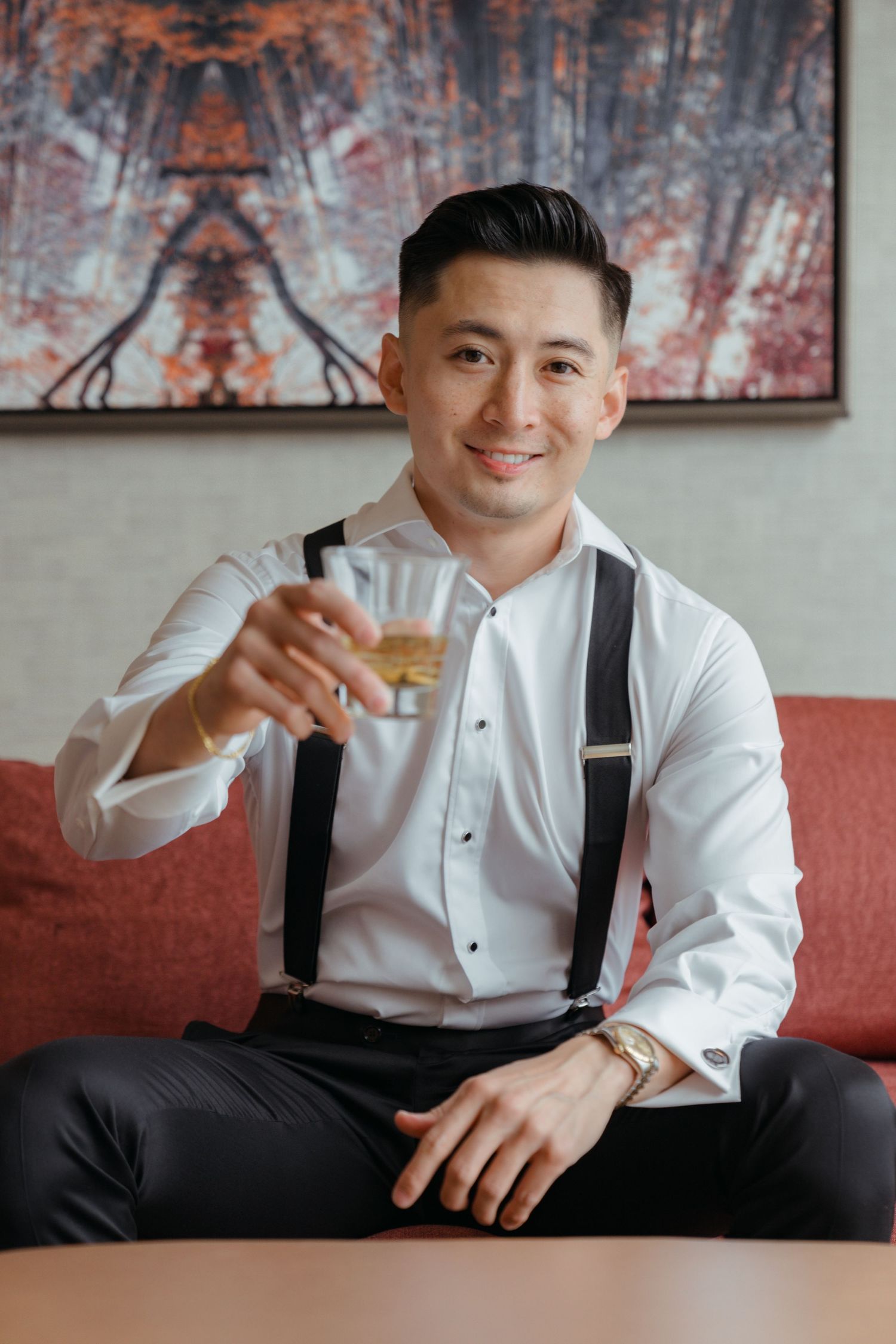 A person in a white dress shirt and suspenders sits casually on a couch, enjoying a drink.