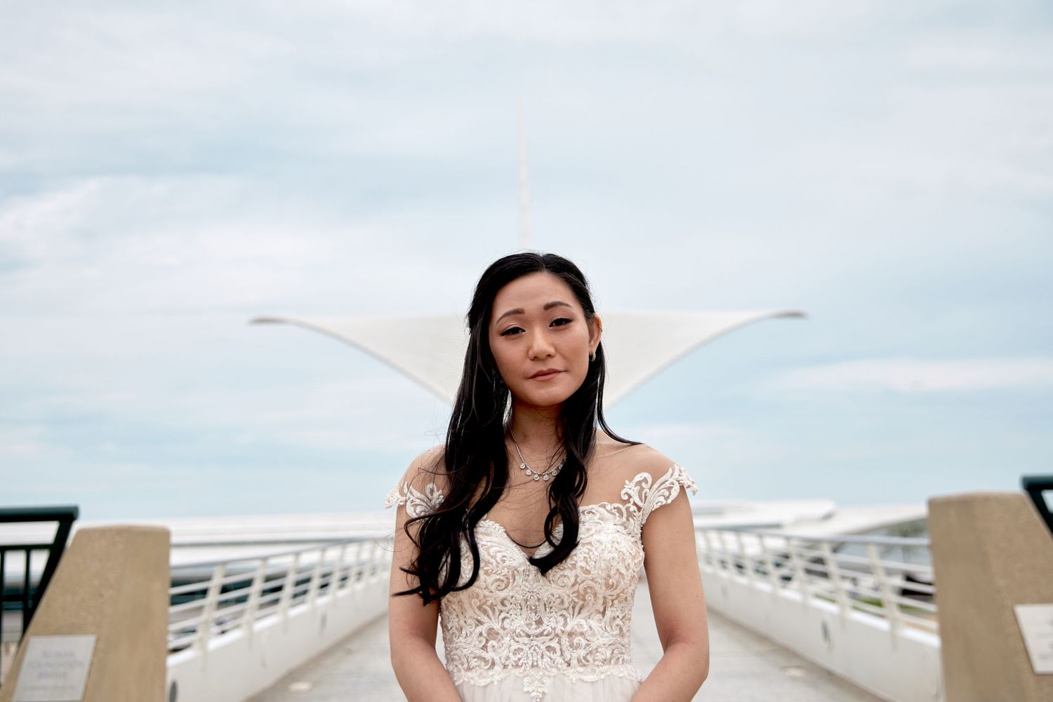Bride in lace wedding dress poses on bridge against cloudy sky.