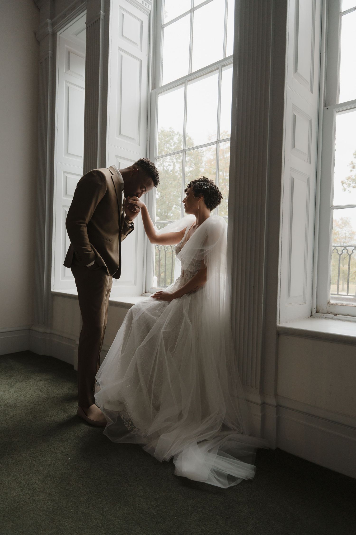 Intimate wedding dress fitting session in sunlit room with tall windows.