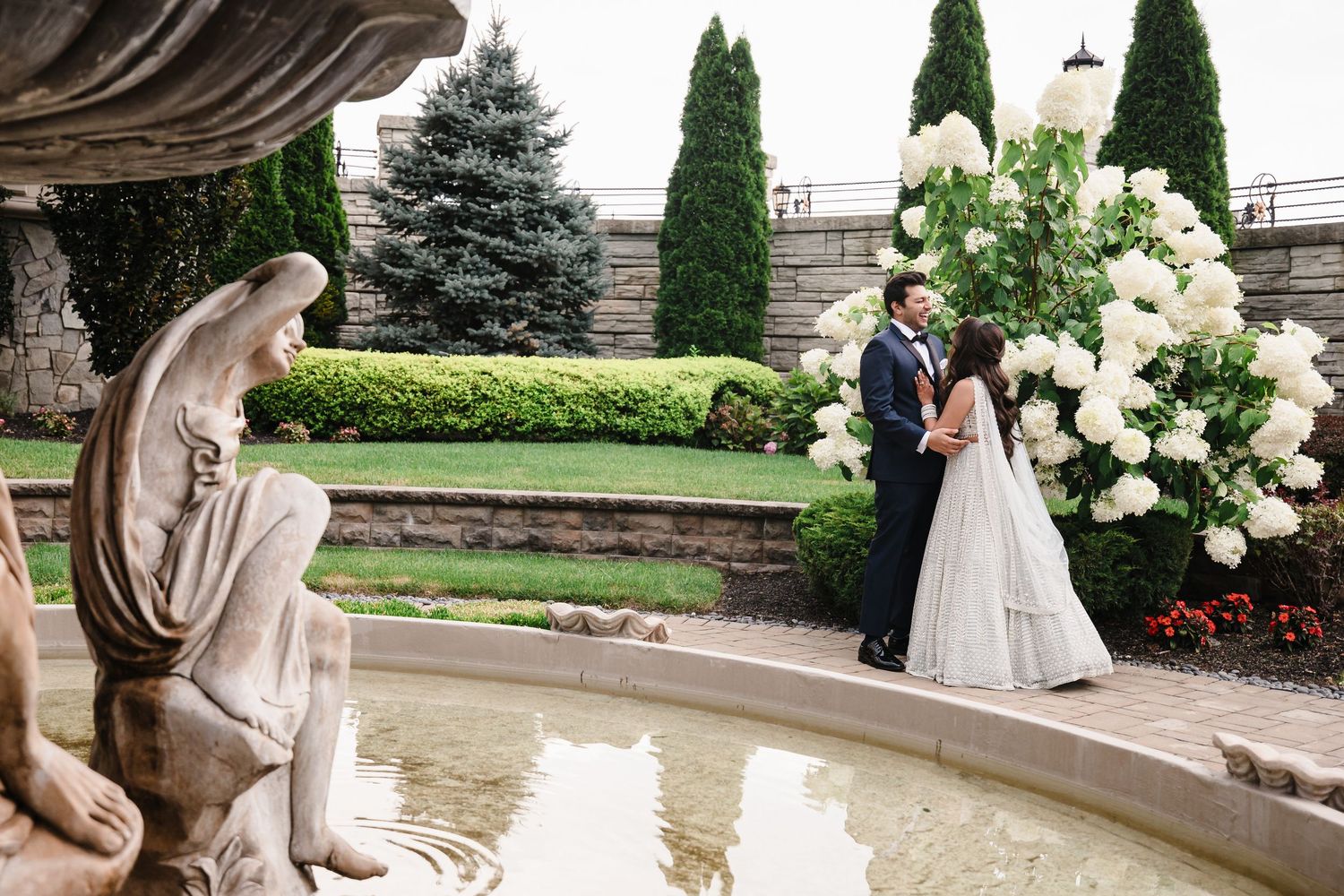 Wedding couple poses near ornate fountain with angel statue and white floral arrangements in garden setting.