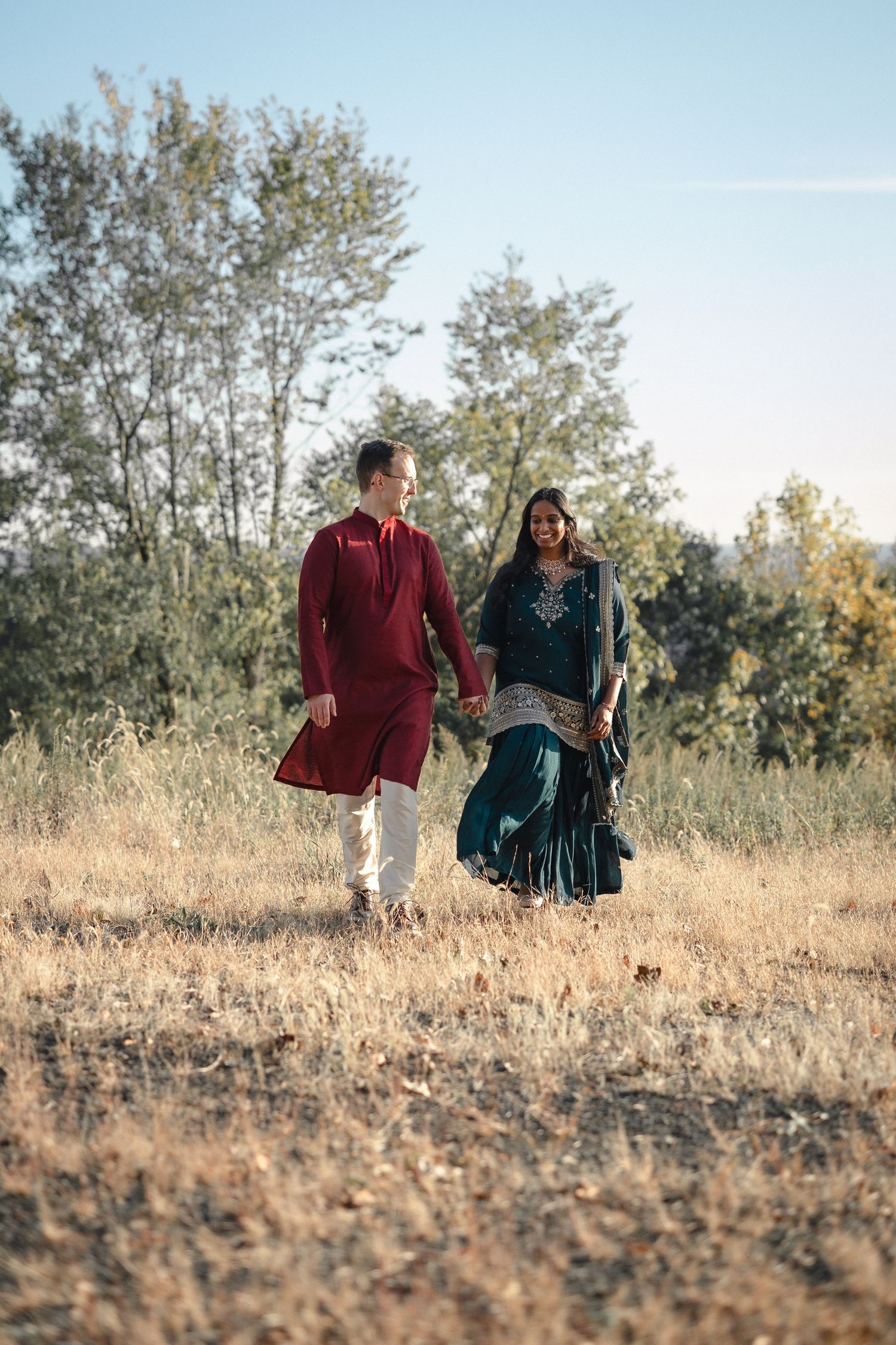 A couple in traditional South Asian attire walks through a sun-lit rural landscape.