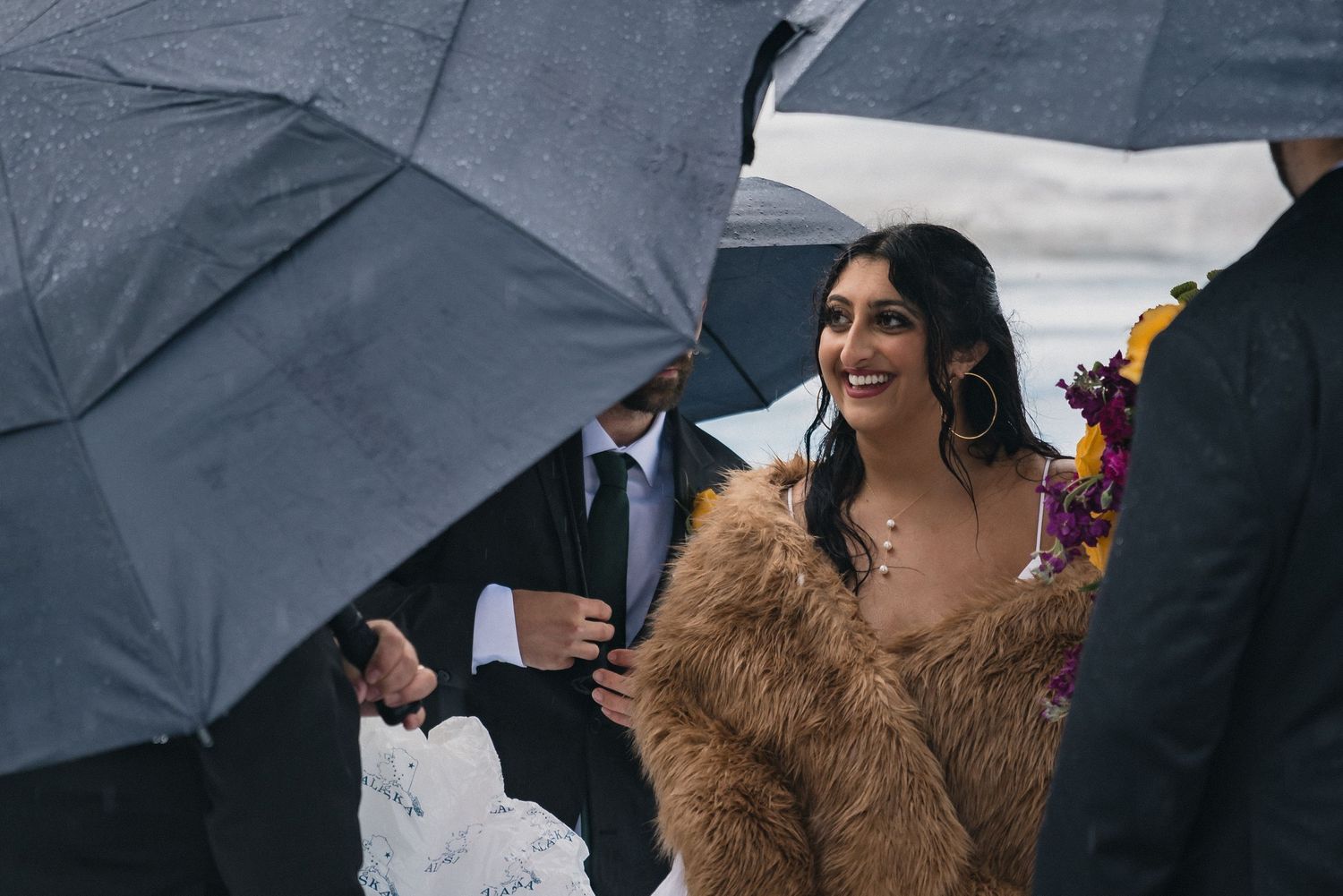 A person in a brown fur coat smiles while standing in the rain under umbrellas at an outdoor event.