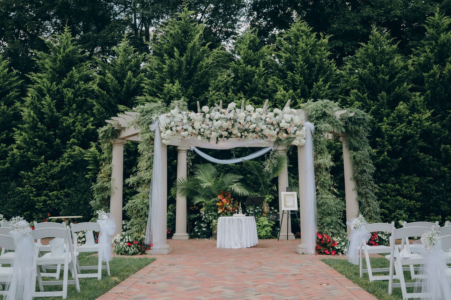 Outdoor wedding ceremony setup with white chairs and floral arch in a garden setting.