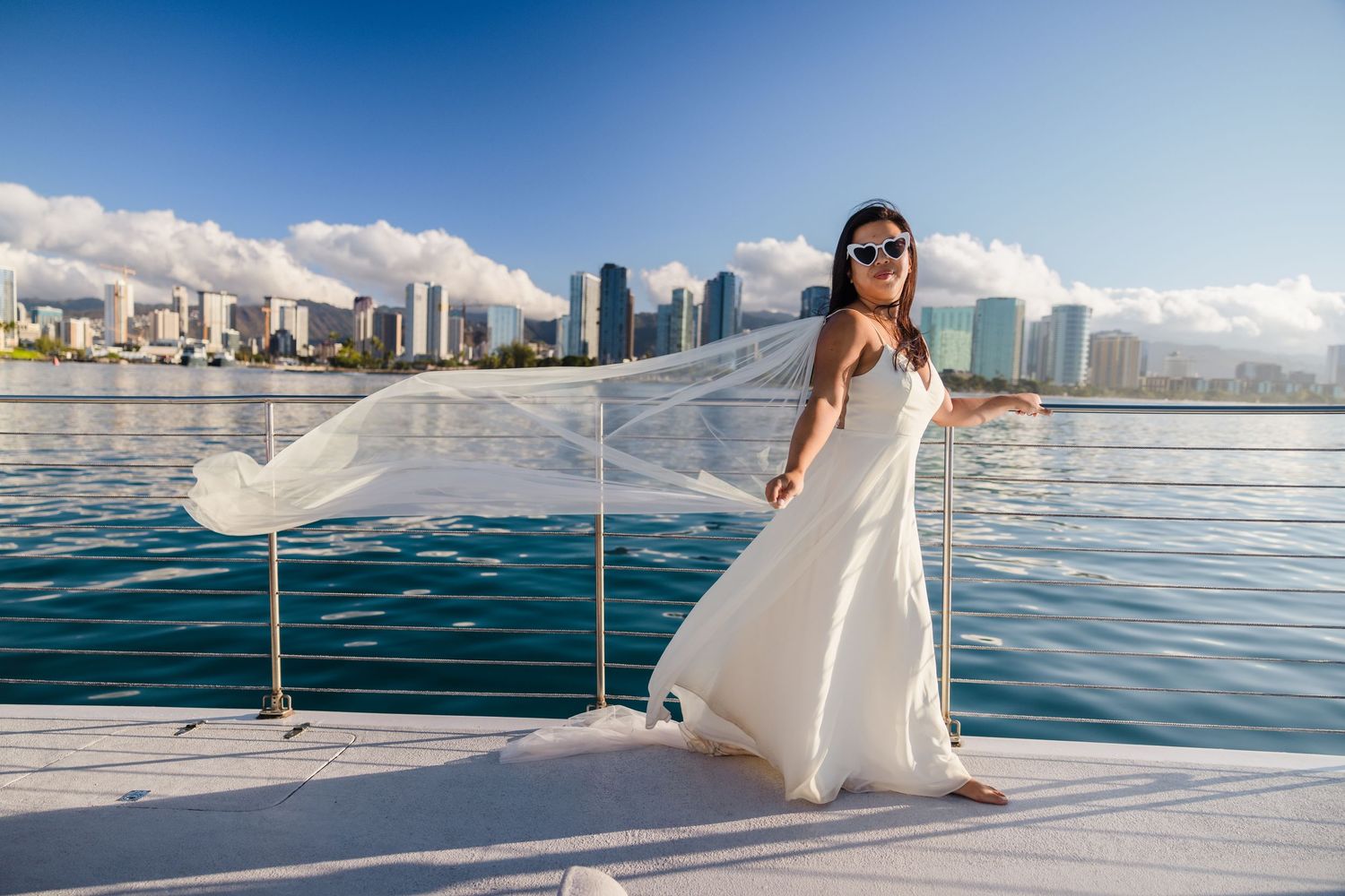 A flowing white wedding dress catches the wind on a boat with city skyline in background.